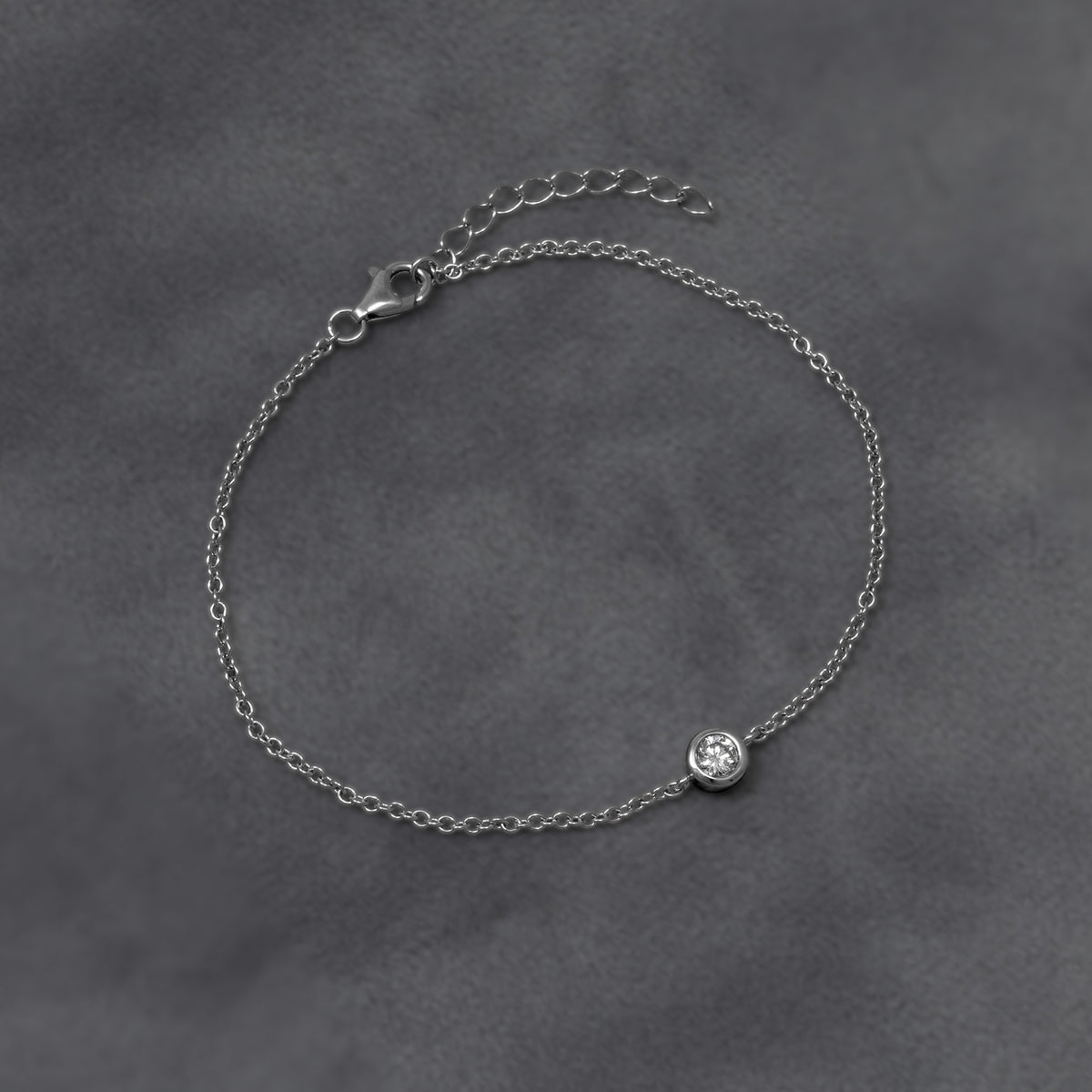 A delicate silver chain bracelet with a bezel-set lab diamond, shown against a grey background.
