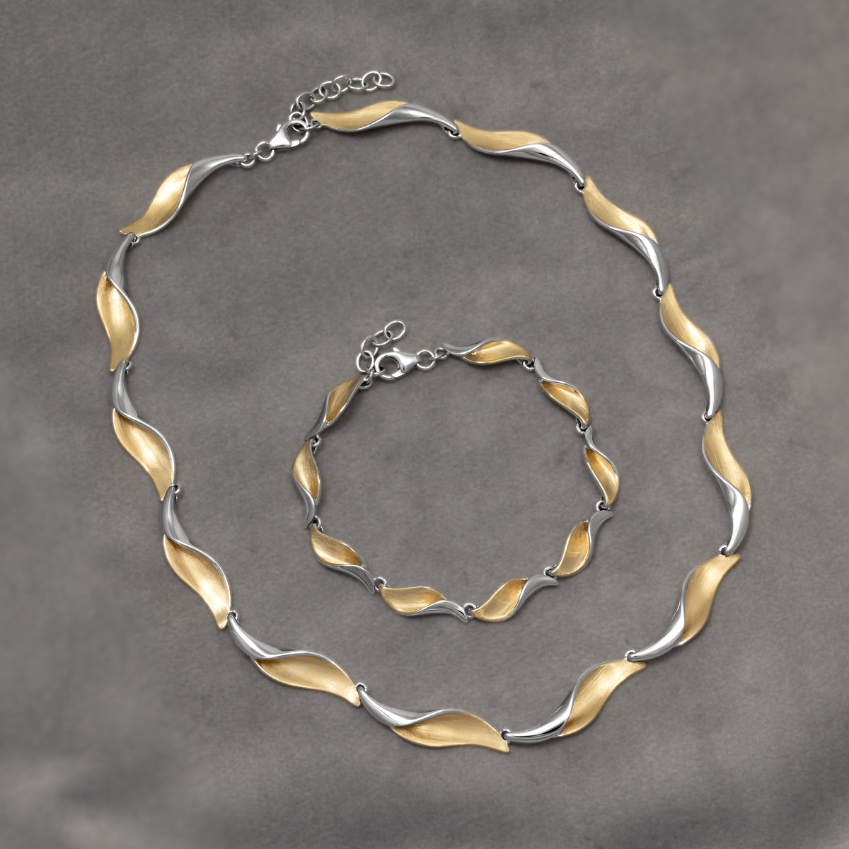 A two-tone link bracelet and necklace set with a wavy, leaf-like design, featuring a satin gold vermeil finish on one side and a polished silver finish on the other, complete with a lobster clasp on a white background.