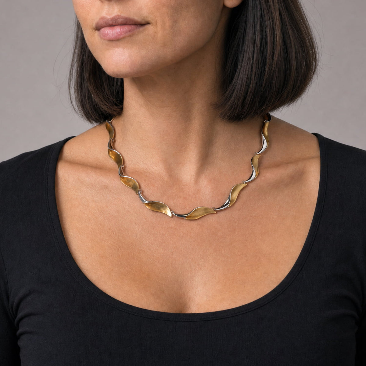 A close-up of a woman wearing a sculptural, wavy two-tone necklace in gold vermeil and silver, paired with a black scoop-neck top against a neutral gray background.