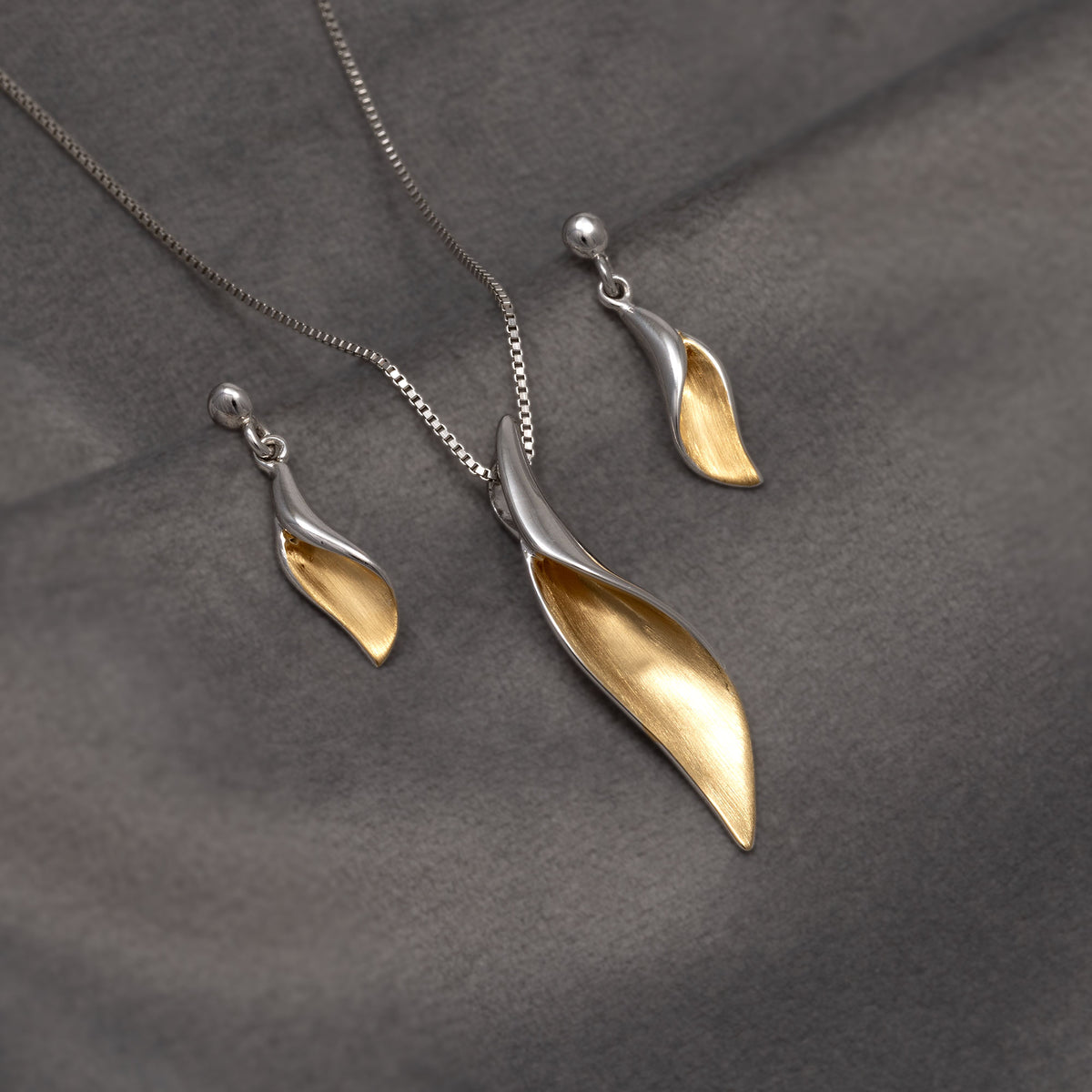 A silver and gold vermeil leaf-shaped jewellery set featuring a pendant and a pair of matching drop earrings displayed on a dark grey textured fabric background.