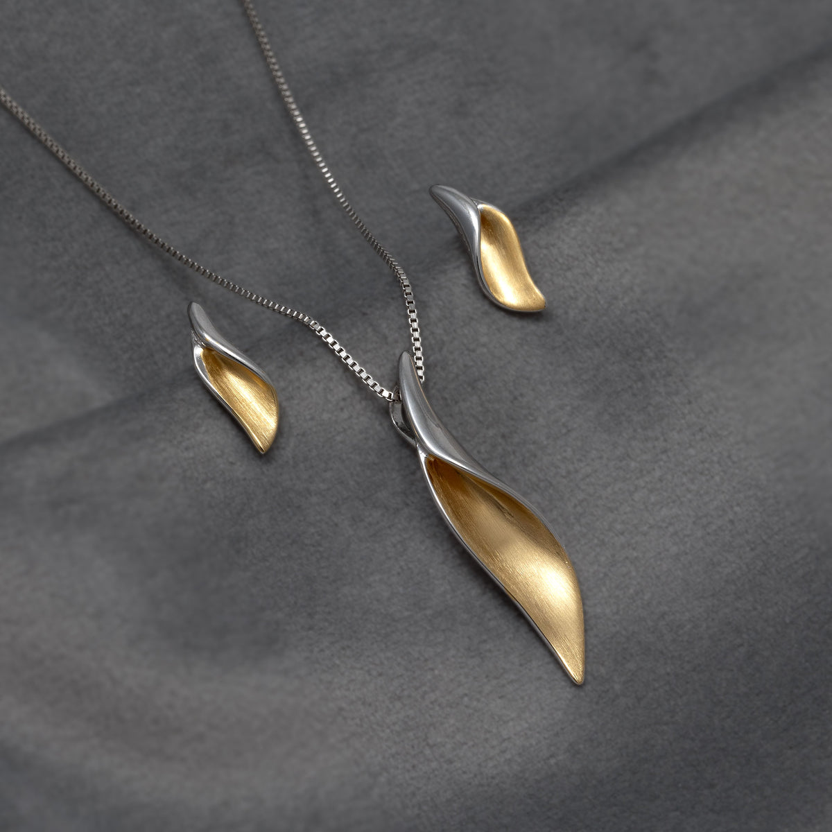 A silver and gold vermeil set in a leaf shape, pendant with matching stud earrings, displayed on a textured dark grey fabric background.
