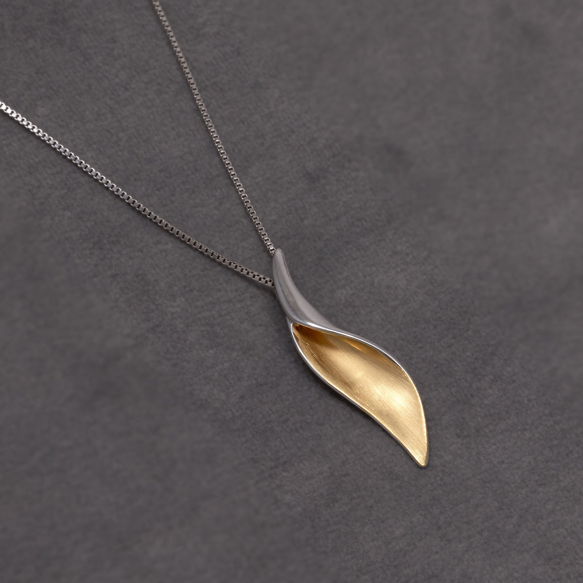 A two-tone leaf-shaped pendant necklace on a delicate silver chain, displayed on a textured dark grey background. The pendant features a polished silver outer edge and a brushed gold vermeil inner surface.