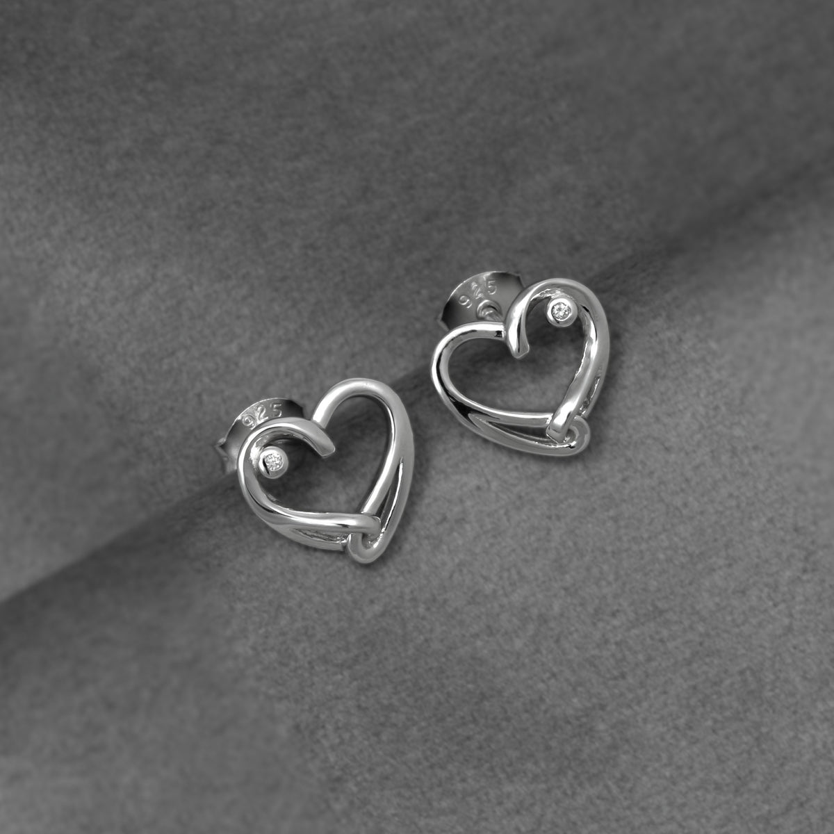 A pair of polished silver heart-shaped stud earrings with lab grown diamond accents, displayed on a textured gray fabric background.