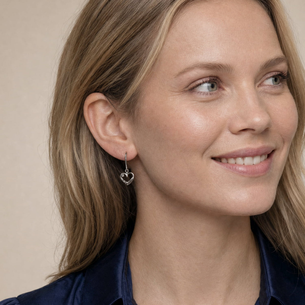 A close-up of a woman with blonde hair wearing a silver drop earring with a heart-shaped dangling charm, set against a soft beige background.