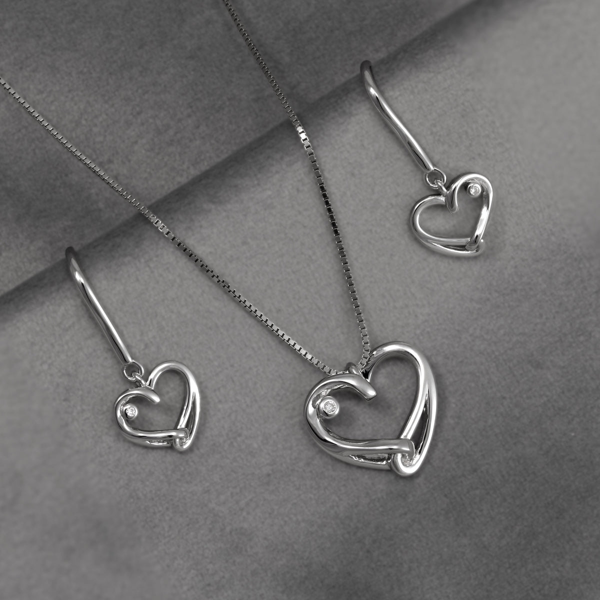 Silver heart pendant and earring set featuring lab grown diamonds, displayed on a textured gray background.