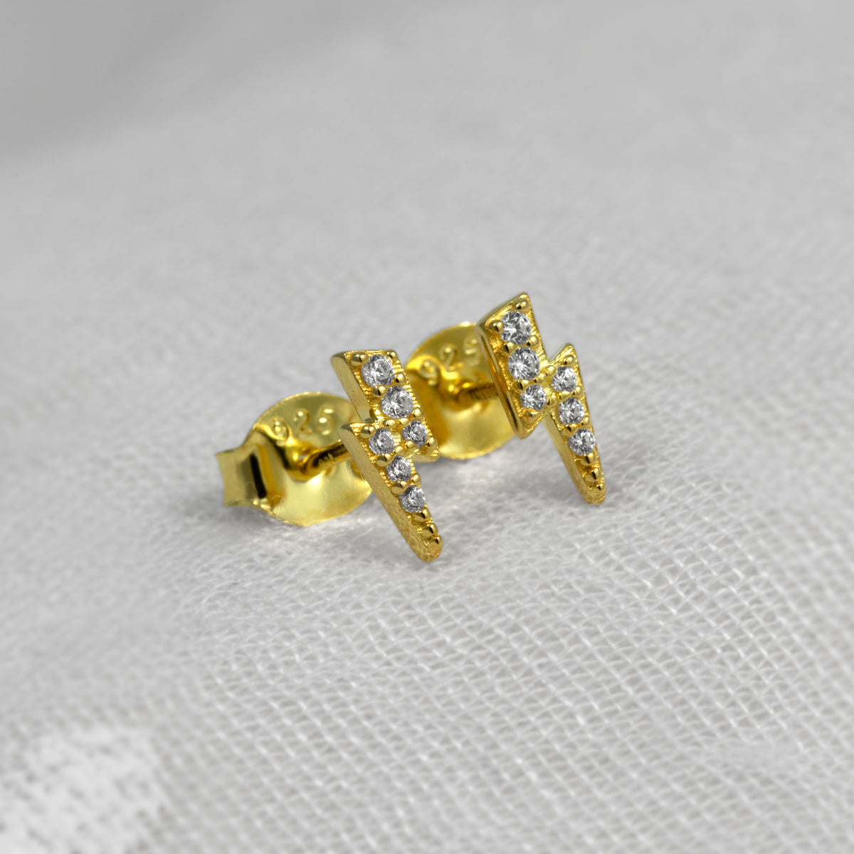 A pair of gold vermeil lightning bolt stud earrings set with small clear cubic zirconia stones, displayed on a textured white fabric background.