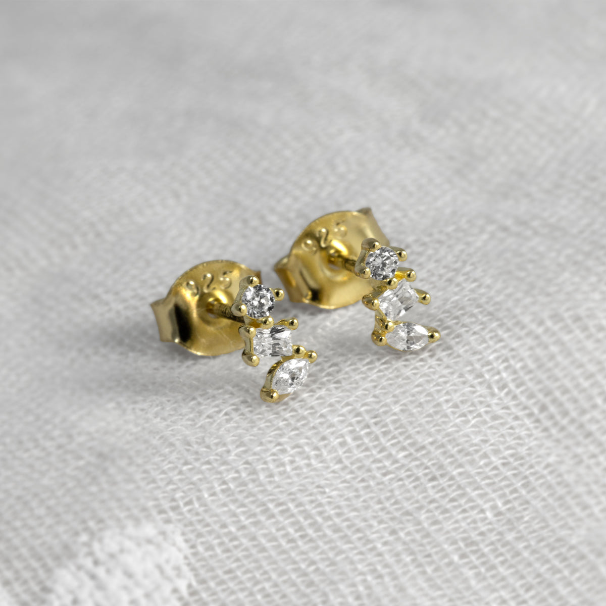A pair of gold vermeil stud earrings with clear marquise-cut and round-cut cubic zirconia, resting on a textured white fabric background.