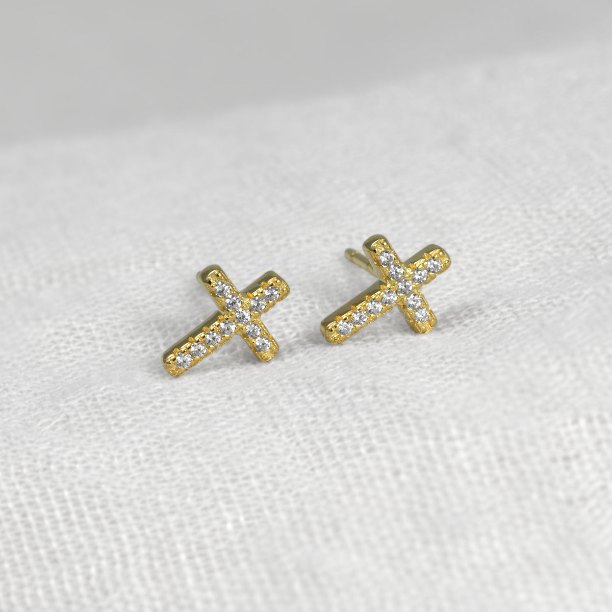 A pair of small gold vermeil cross stud earrings set with clear pavé gemstones, resting on a textured white fabric background.