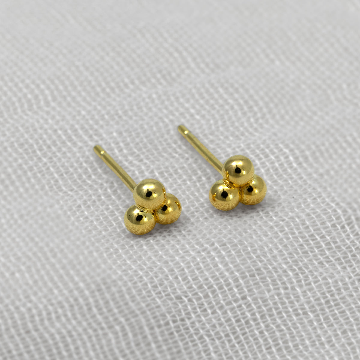 A pair of small gold vermeil stud earrings with three polished spherical beads clustered on each post, photographed on a textured white fabric background.