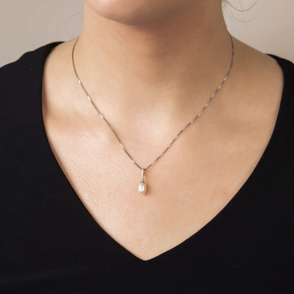 A close-up of a delicate silver chain necklace with a pavé-set oval pearl pendant worn on a woman’s neckline against a black top, highlighting fine designer craftsmanship.