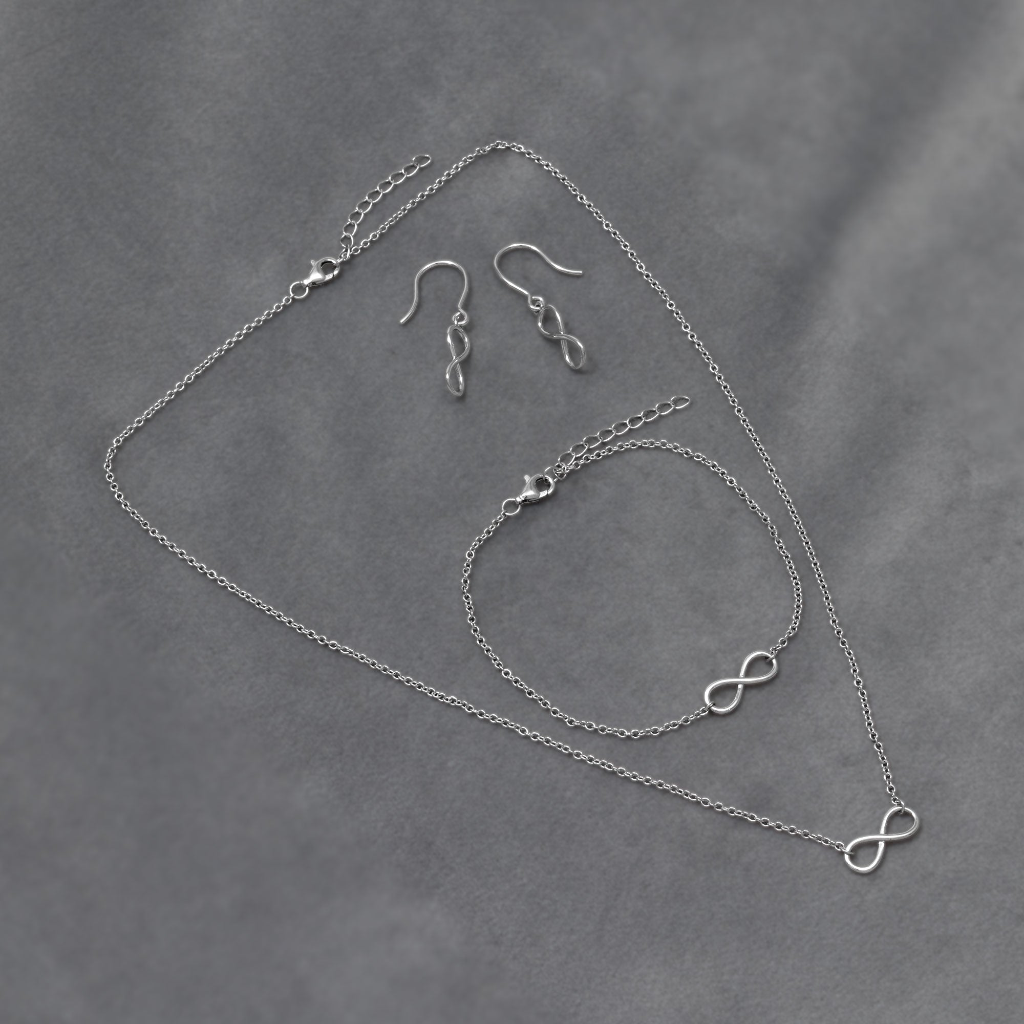 A delicate sterling silver necklace, bracelet and drop earrings featuring a simple infinity symbol, embodying elegant and timeless design. Placed on a grey velvet cloth.