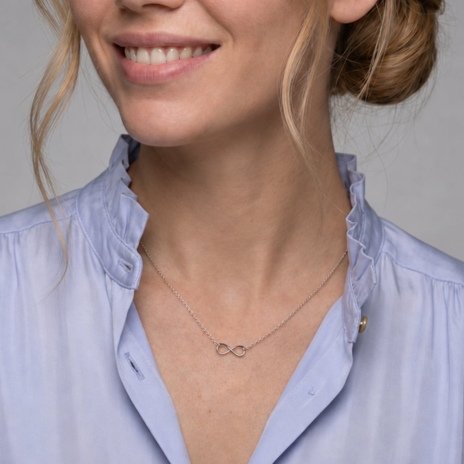 A close-up of a woman wearing a delicate silver infinity necklace, paired with a light blue ruffled-collar blouse.