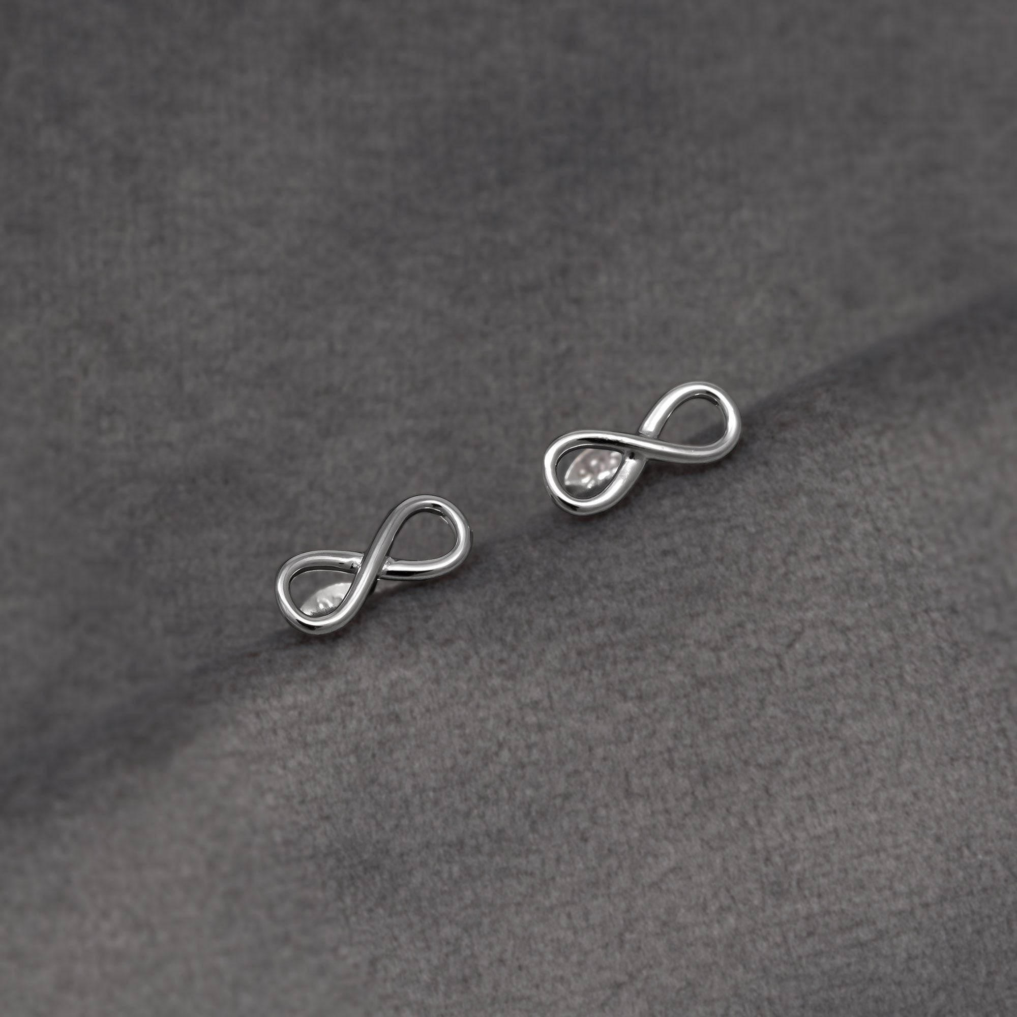 Pair of minimalist sterling silver stud earrings shaped as smooth infinity symbols with secure butterfly backs, embodying elegant and contemporary silver jewellery design. Placed on a grey velvet cloth.