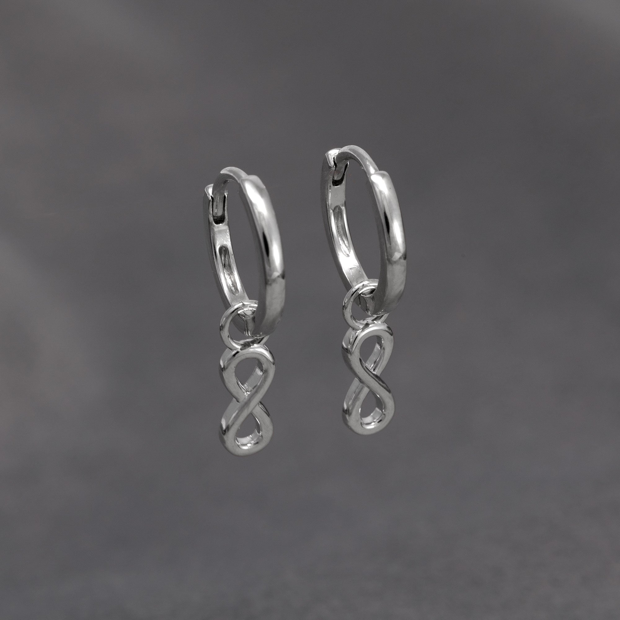 A pair of sleek sterling silver hoop earrings featuring small, polished infinity-shaped charms hanging delicately from each hoop, embodying minimalist elegance. Placed on a grey velvet cloth.