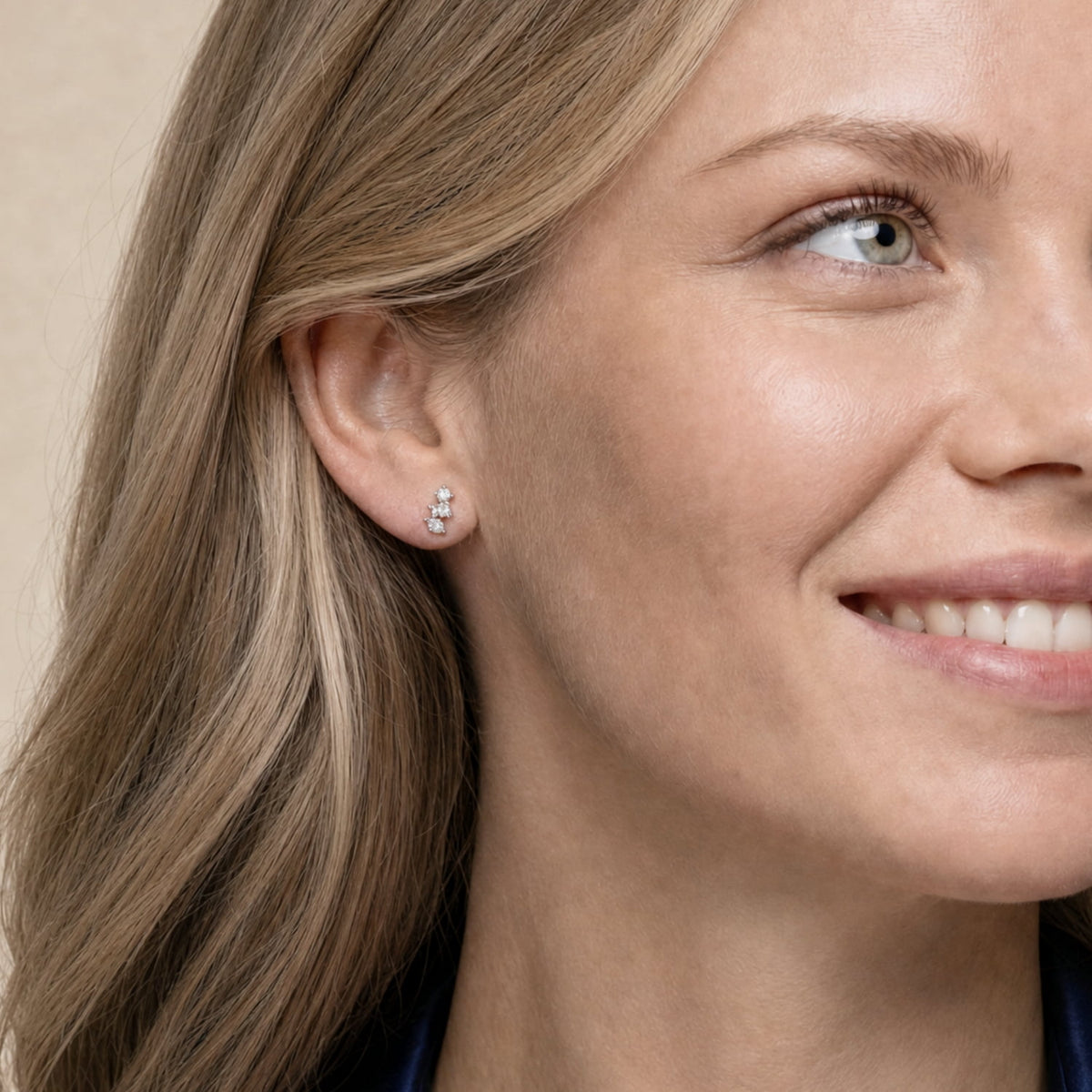 A close-up of a woman's ear wearing a small cluster stud earring with clear cubic zirconia in a silver setting, against a soft beige background.