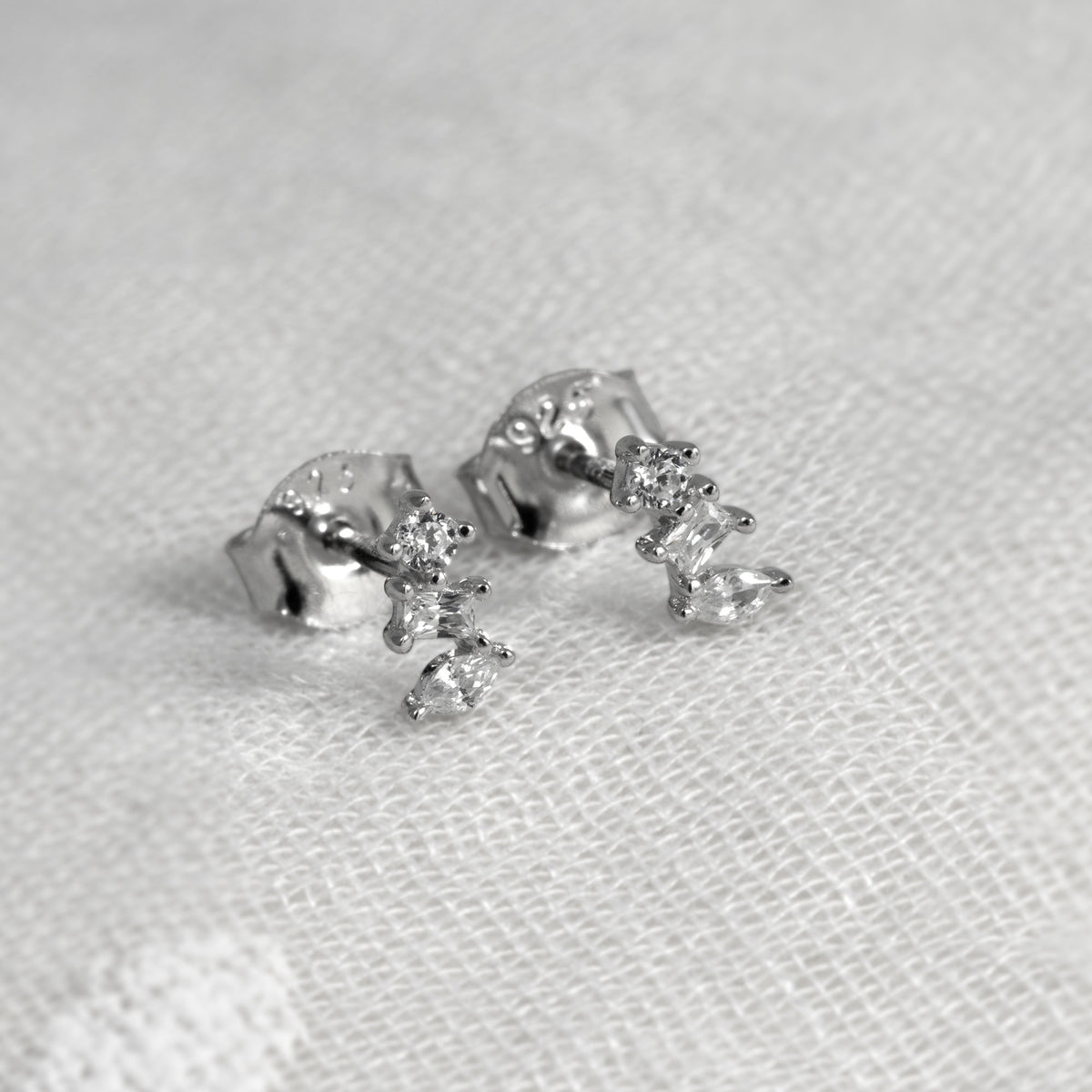 A pair of silver stud earrings with small clear cubic zirconia, resting on a textured white fabric background.