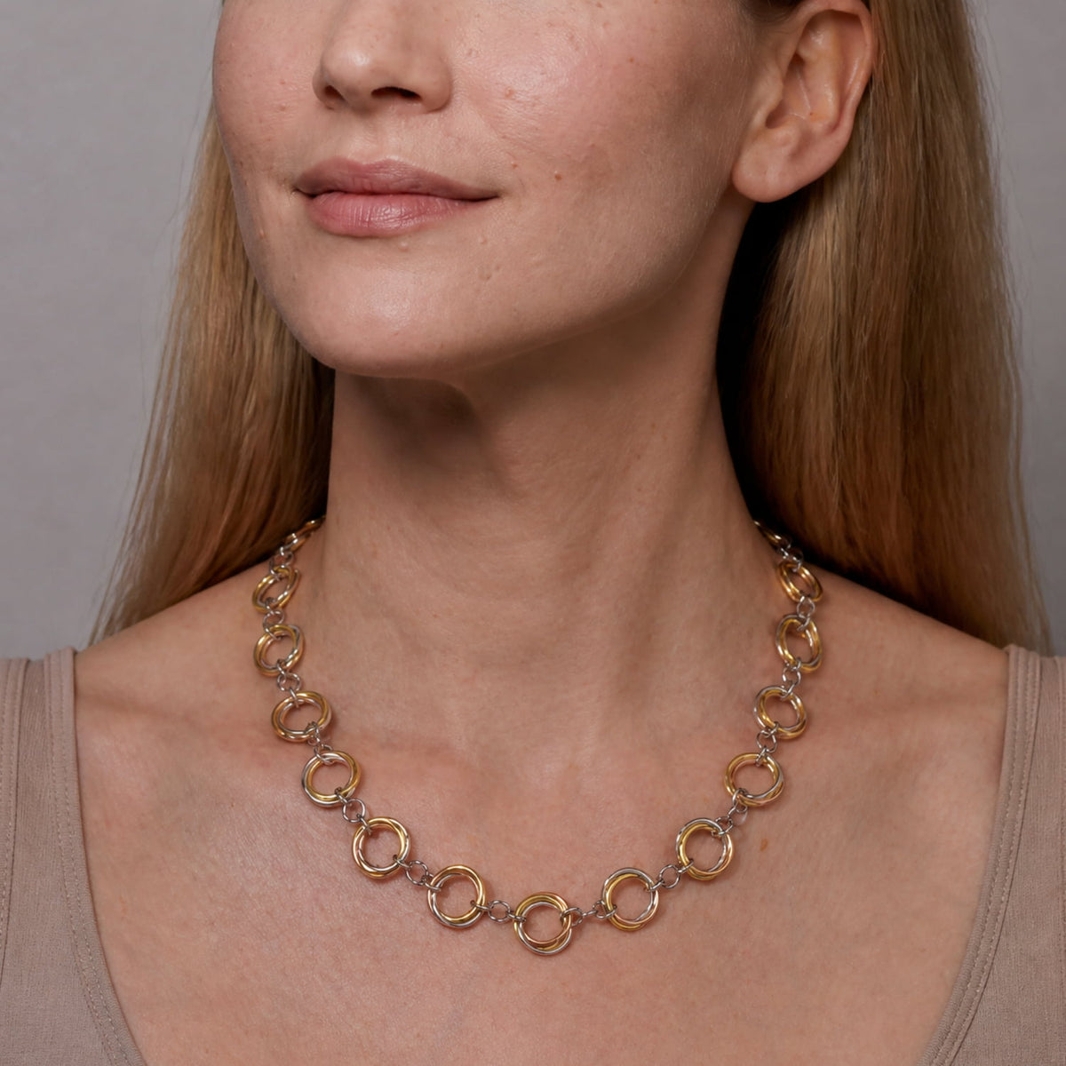 A close-up of a woman wearing a three tone ring-link necklace, paired with a beige top against a warm beige background.