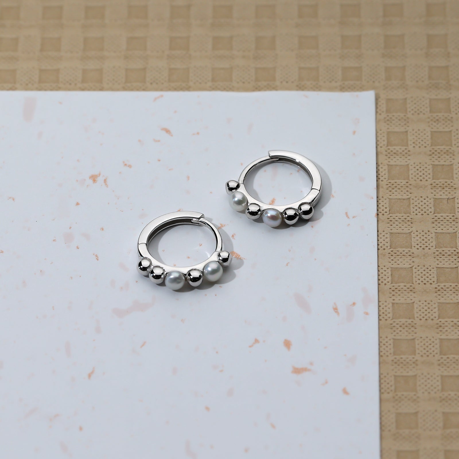 A pair of sterling silver hoop earrings featuring a row of alternating smooth beads and lustrous pearls, displayed on a white and beige textured surface.