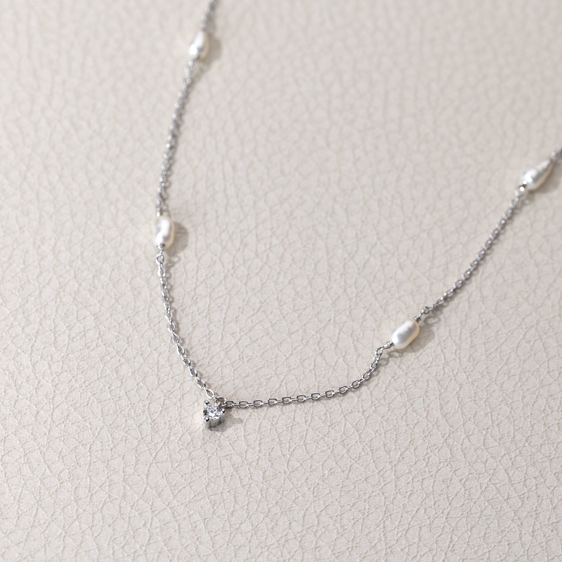 A delicate sterling silver chain necklace featuring spaced freshwater pearls and a small cubic zirconia charm resting on a textured neutral background.