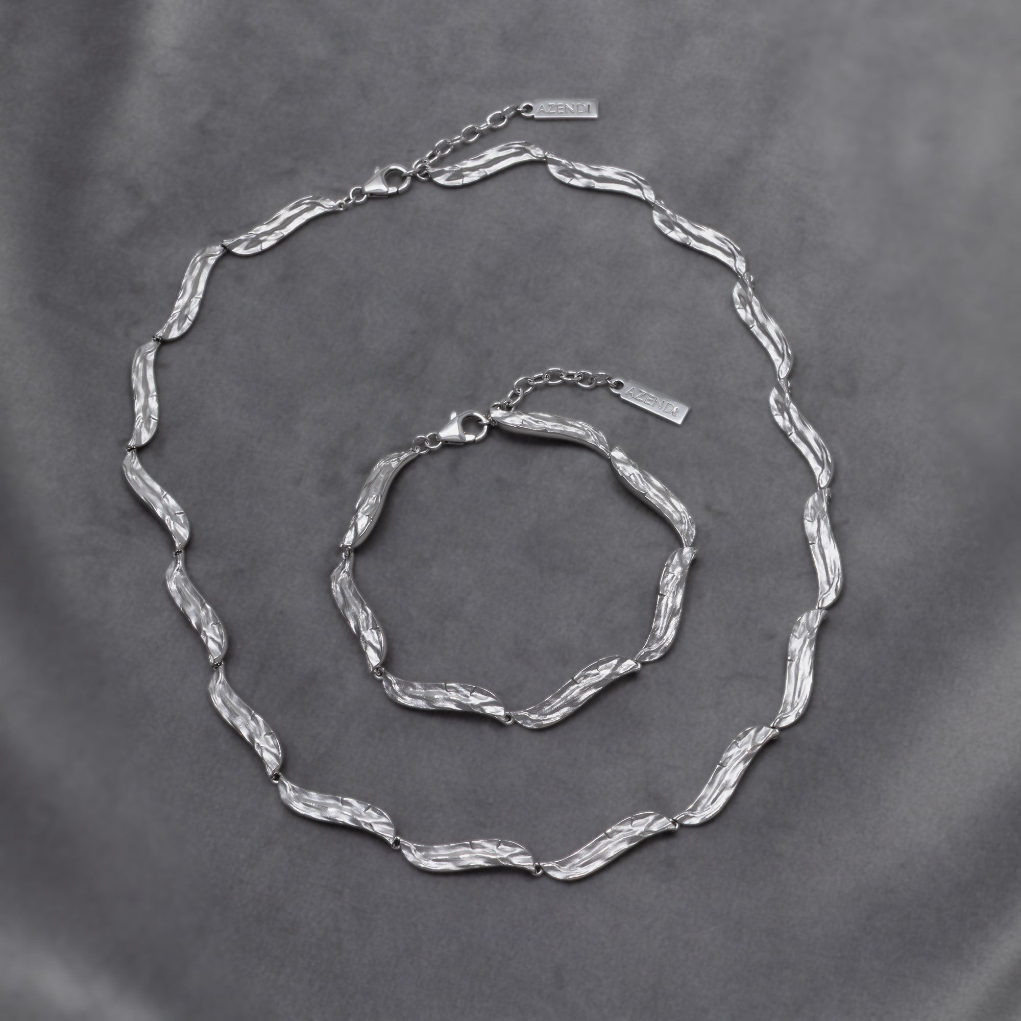 Sculptural sterling silver necklace and matching bracelet featuring textured, undulating links with adjustable chain closures, showcasing contemporary British silver jewellery design. Placed on a grey velvet cloth.