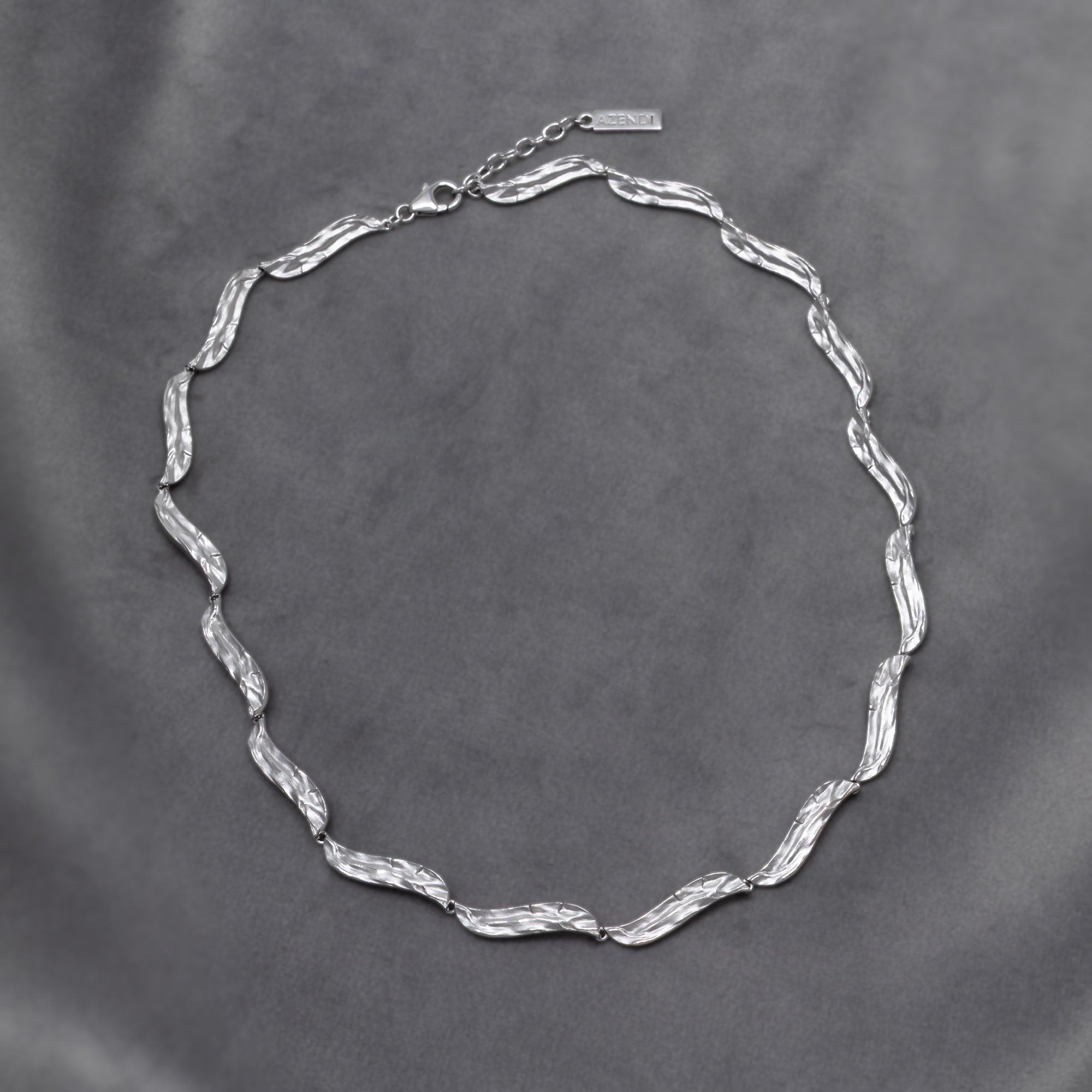 Sculptural sterling silver necklace featuring textured, undulating links with adjustable chain closures, showcasing contemporary British silver jewellery design. Placed on a grey velvet cloth.