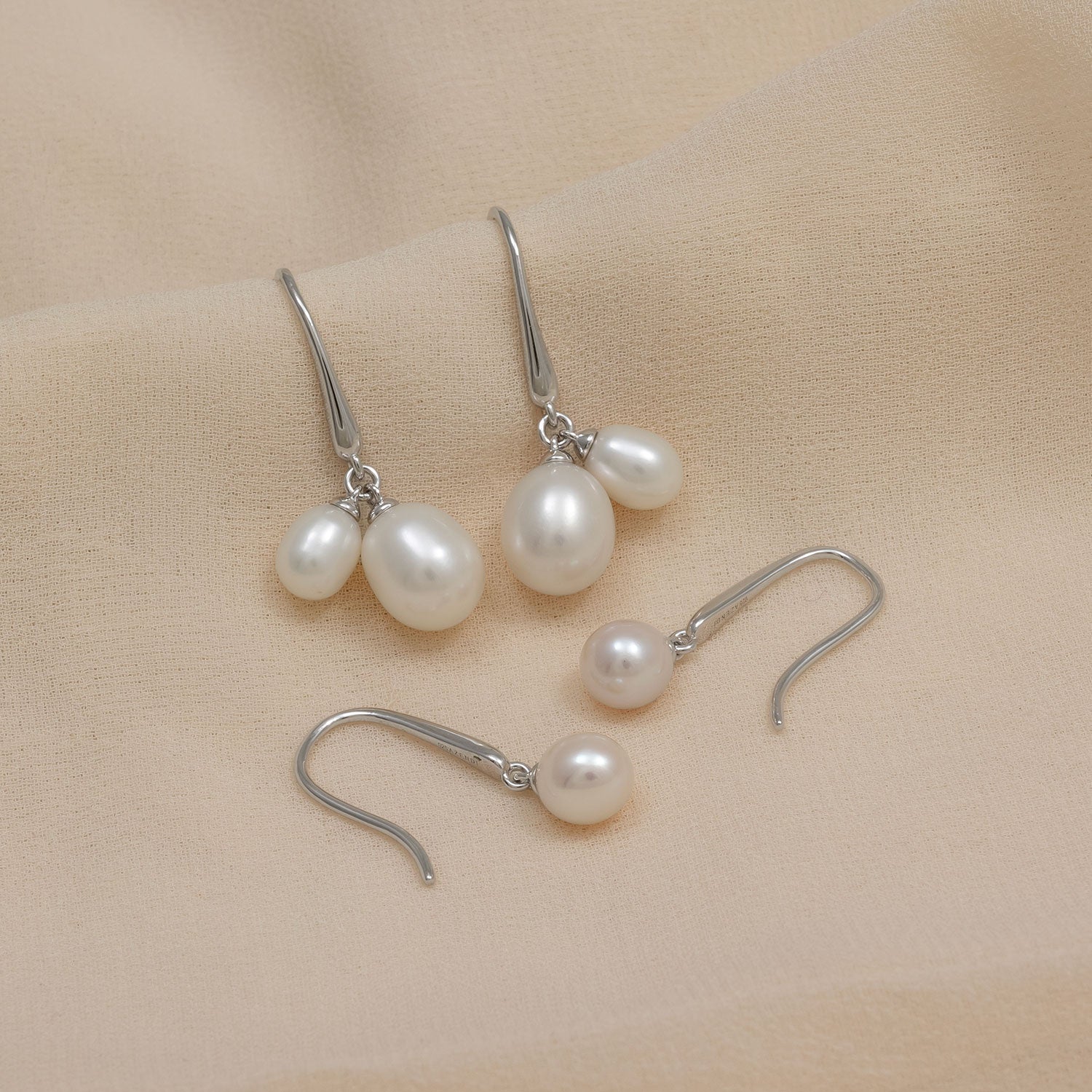 Two pairs of sterling silver hook earrings featuring lustrous white pearls in varied sizes, displayed on a soft beige fabric background.