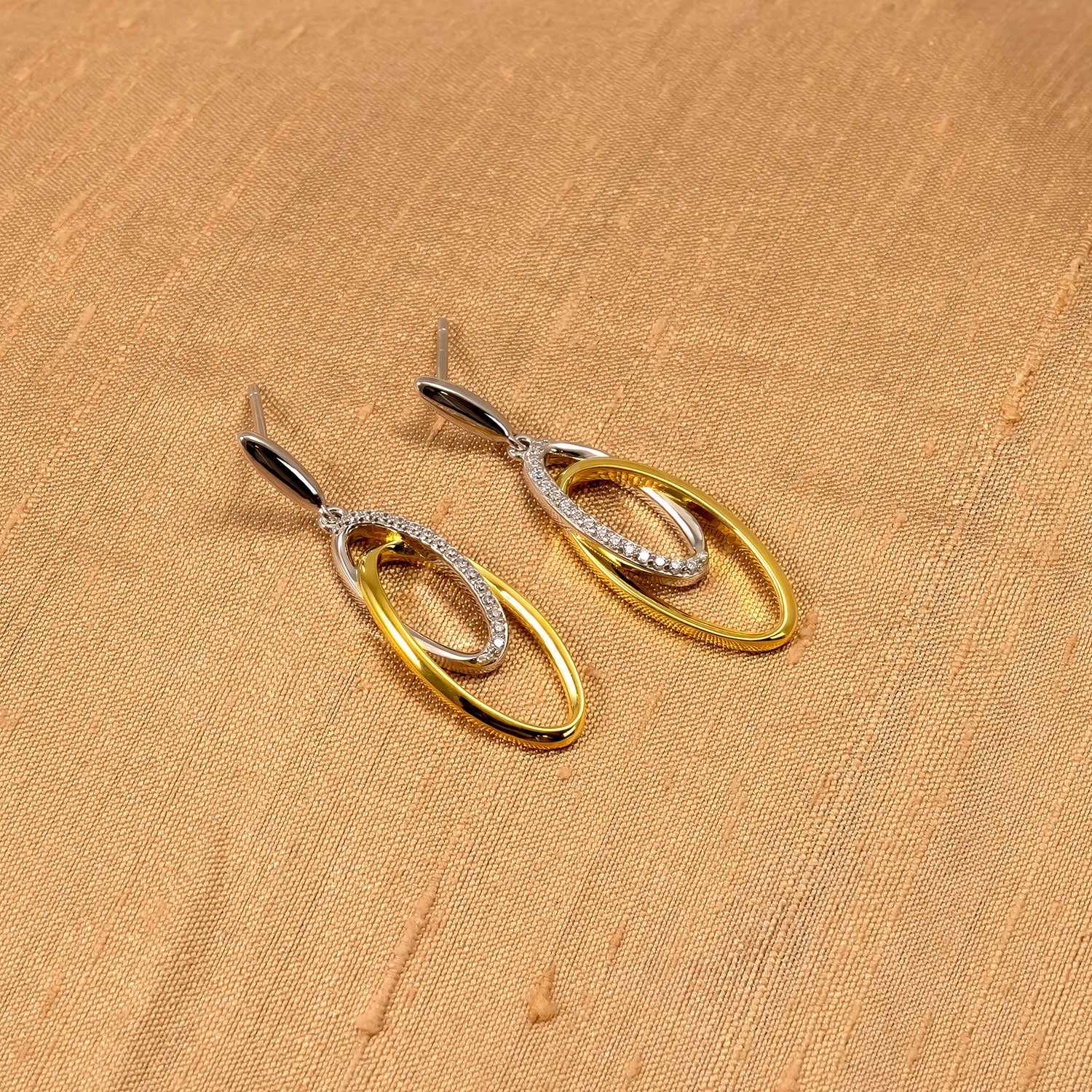 Pair of drop earrings featuring interlocking oval shapes in sterling silver and gold vermeil with a polished finish, displayed on a warm textured fabric background.