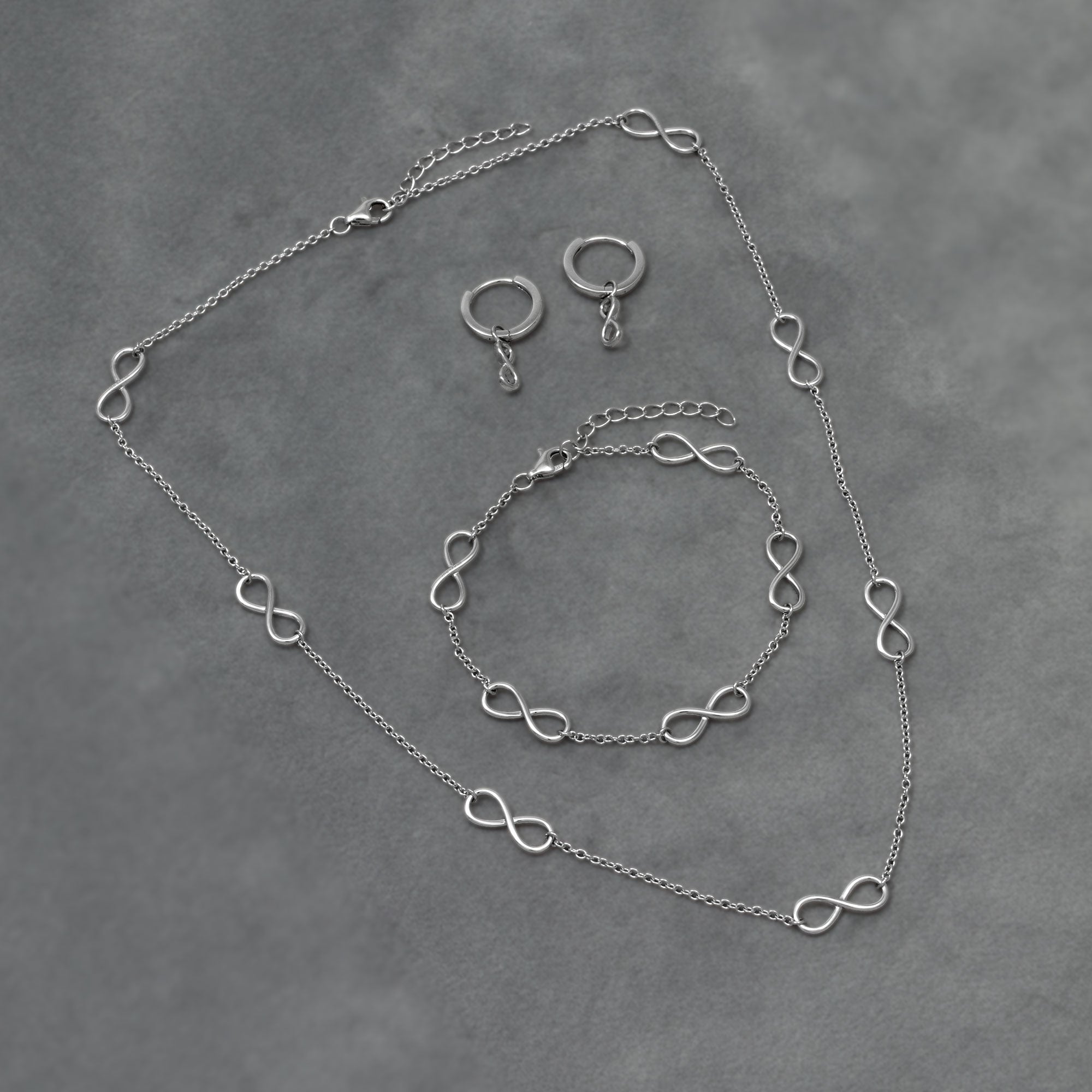 Delicate sterling silver necklace, bracelet and drop earring featuring multiple small infinity symbols, showcasing a minimalist and elegant design. Placed on a grey velvet cloth.