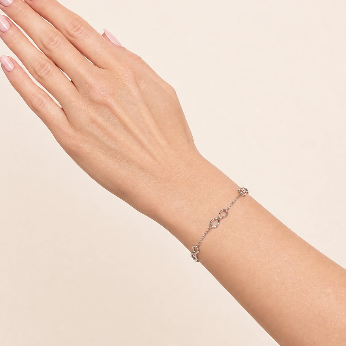 A delicate silver bracelet with multiple infinity symbols, shown worn on a woman's wrist against a soft beige background.