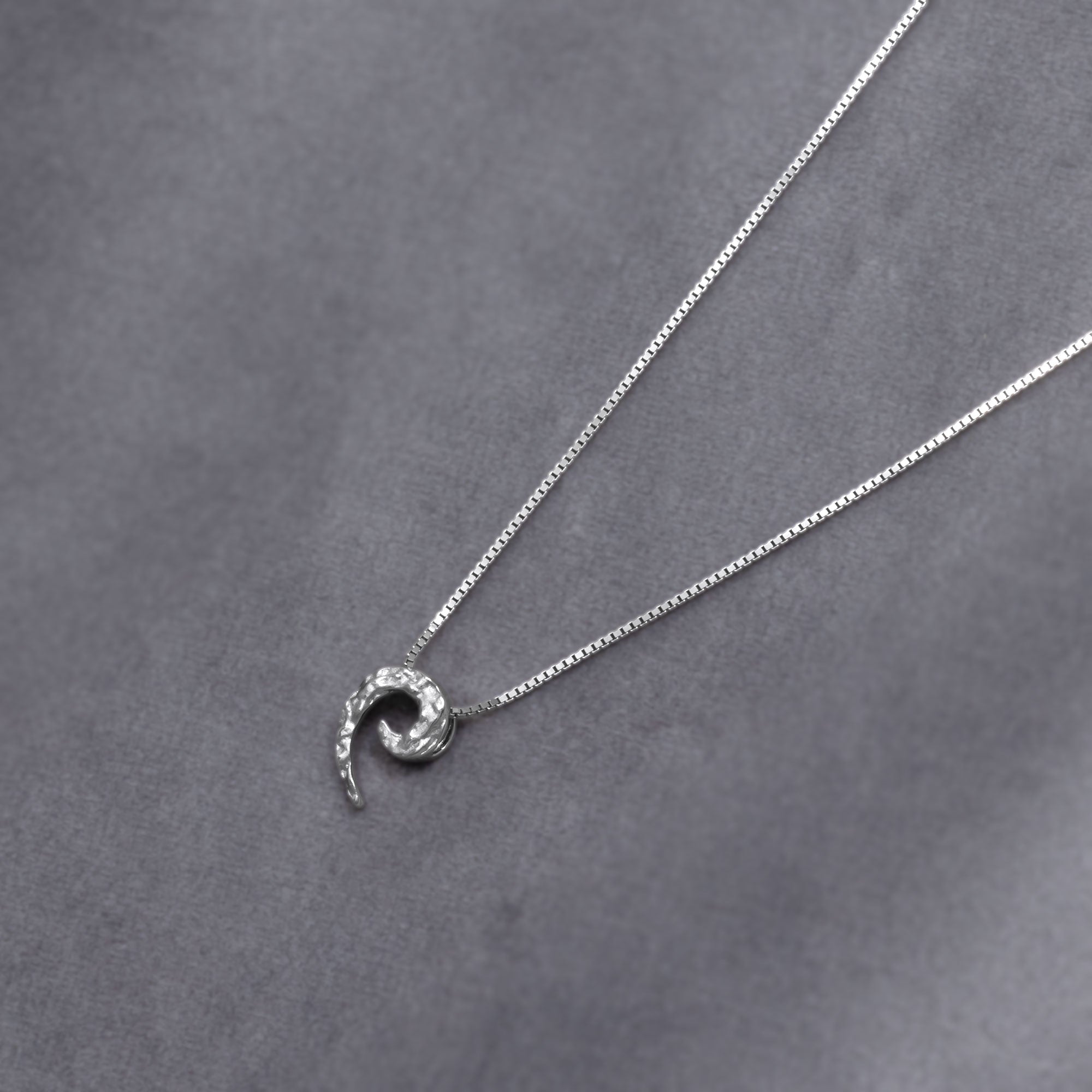 Sterling silver pendant with a textured curling horn design suspended on a delicate box chain, exemplifying sculptural elegance in contemporary silver jewellery. Placed on a grey velvet cloth.
