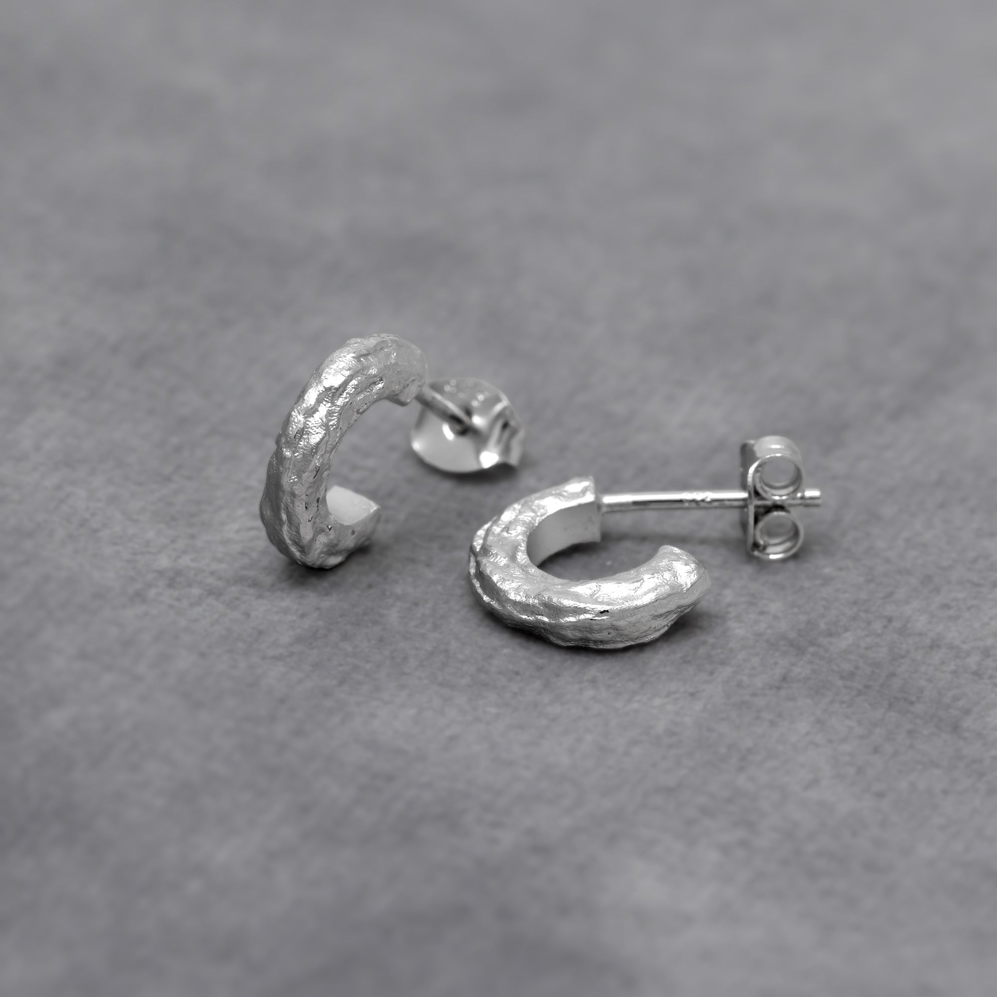 Pair of textured sterling silver half hoop earrings with a sculptural, hammered finish, offering a contemporary and elegant design in fine silver jewellery by Azendi. Placed on a grey velvet cloth.