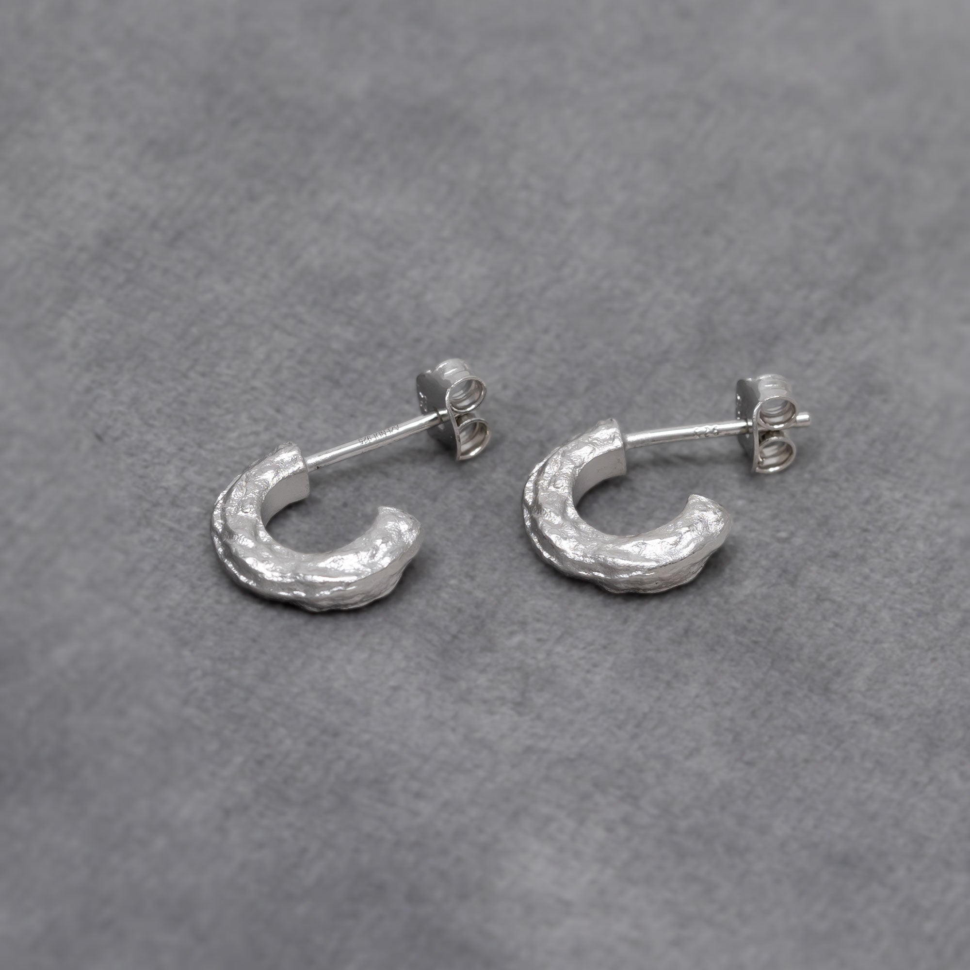 Pair of textured sterling silver half hoop earrings with a sculptural, hammered finish, offering a contemporary and elegant design in fine silver jewellery by Azendi. Placed on a grey velvet cloth.