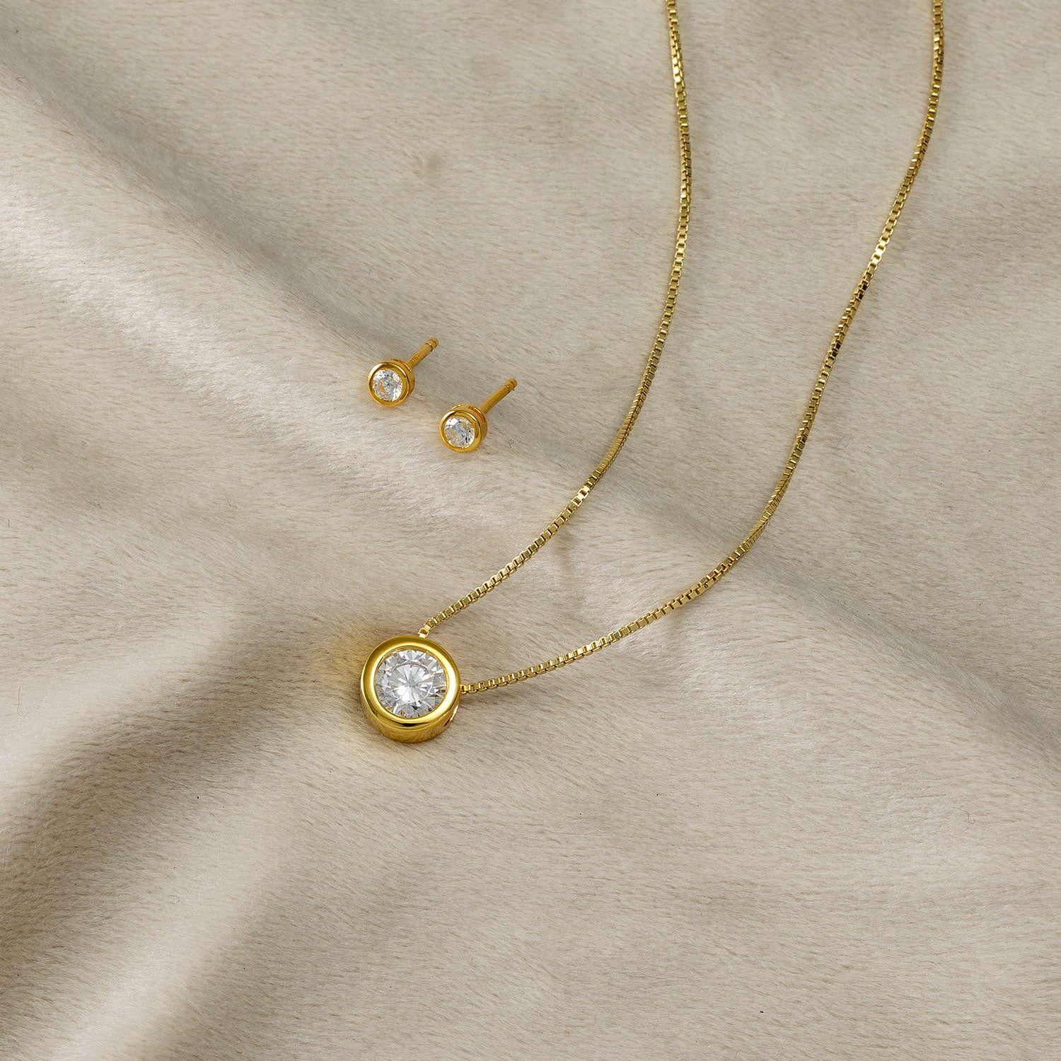 Gold vermeil stud earrings with brilliant cut clear stones paired with a matching pendant necklace set against a soft beige fabric background.