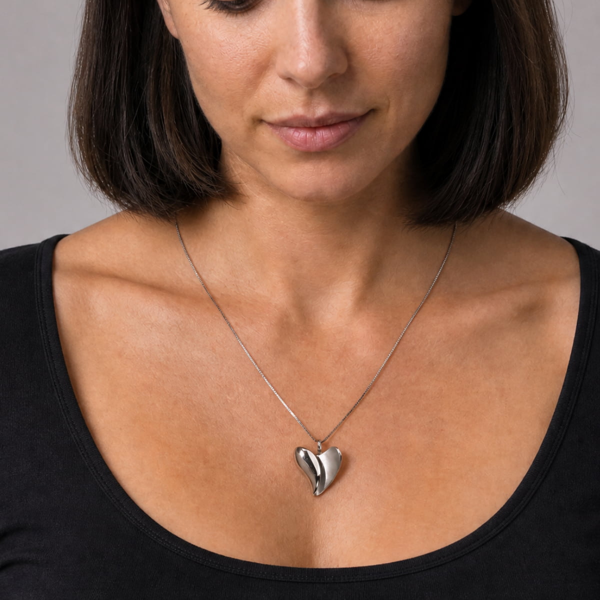 A close-up of a woman wearing a silver heart pendant necklace on a delicate chain, paired with a black scoop-neck top against a neutral gray background.