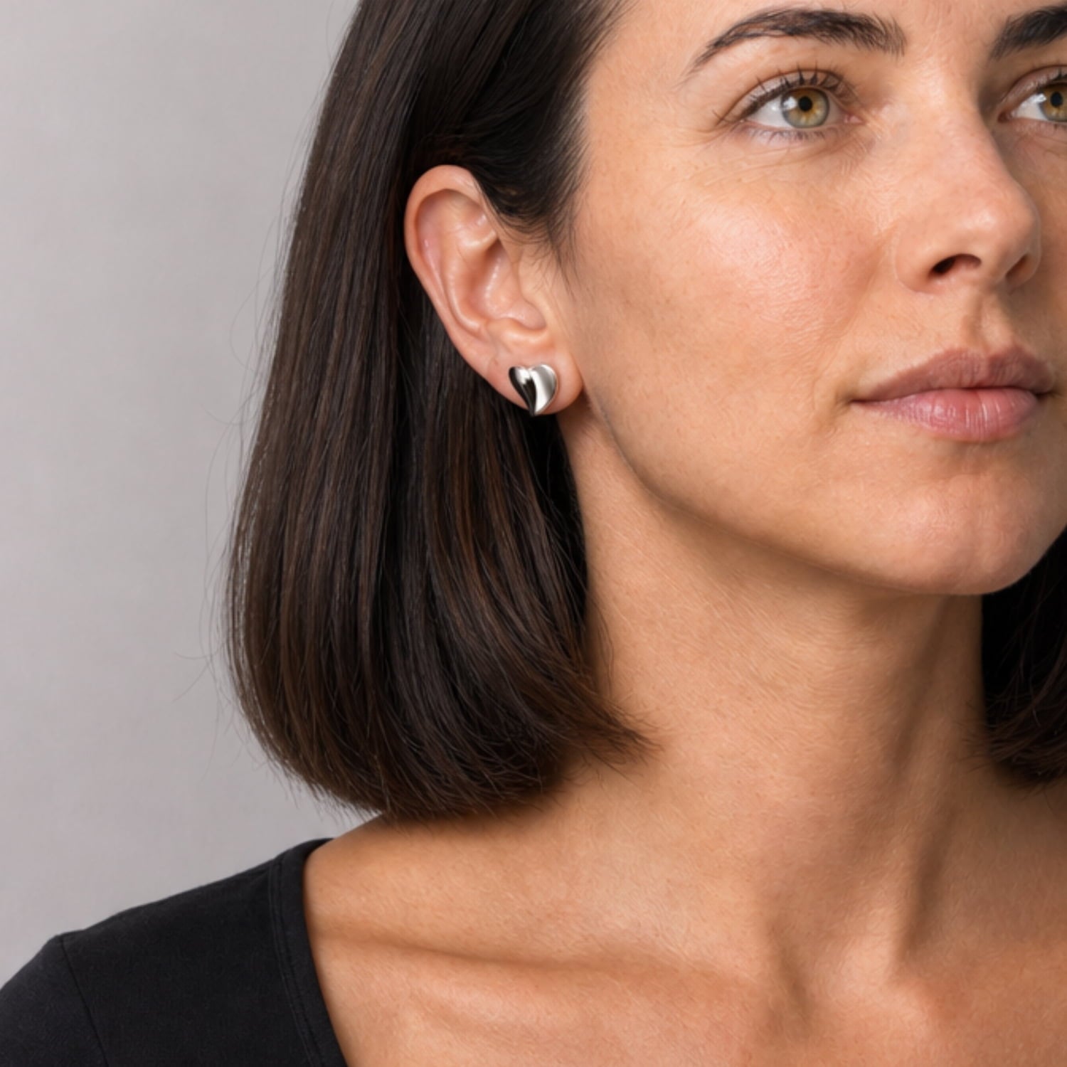 Woman wears a pair of sterling silver stud earrings crafted in a curling heart shape, featuring smooth, flowing contours that highlight a modern sculptural aesthetic.