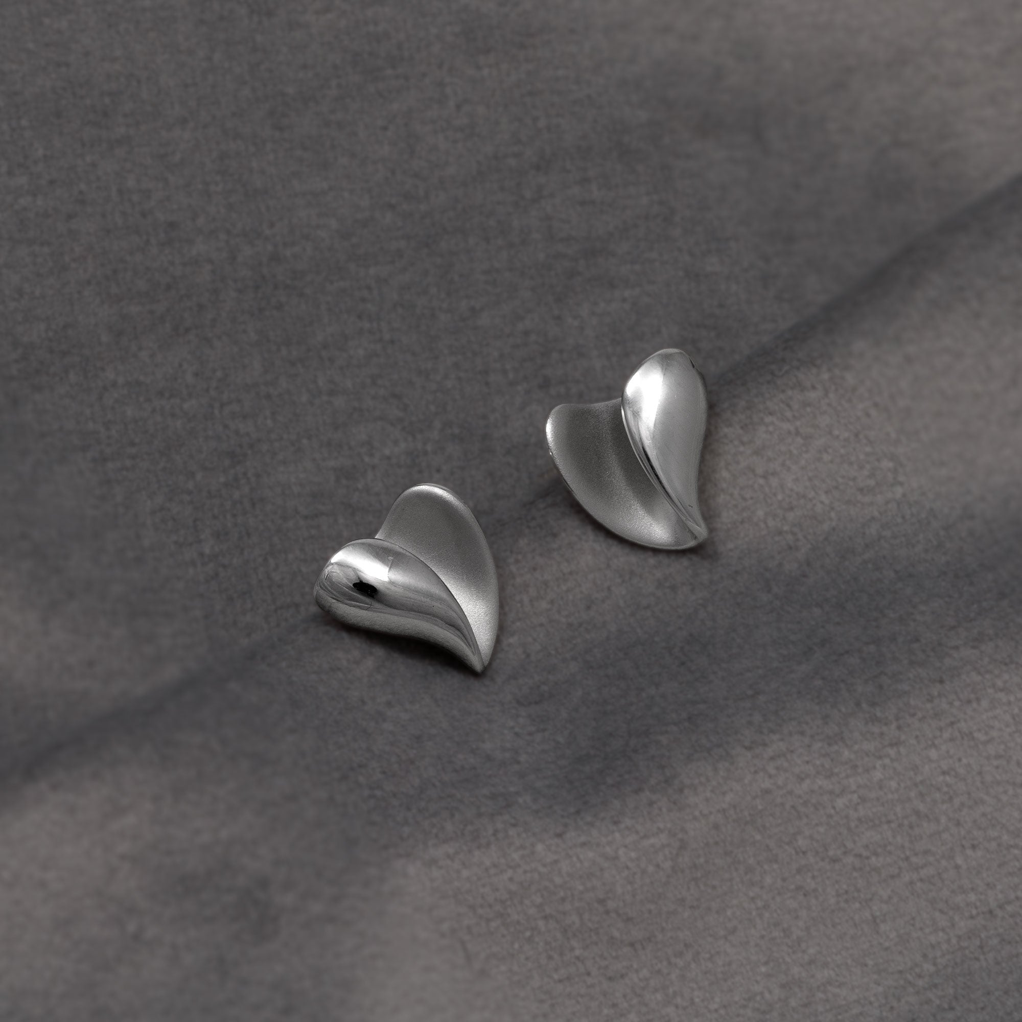 A pair of polished silver heart-shaped stud earrings resting on a textured dark grey fabric background.