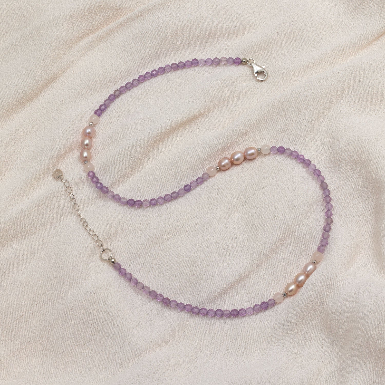 Delicate sterling silver necklace featuring faceted amethyst beads interspersed with clusters of lustrous freshwater pearls, laid out on a soft beige fabric background.