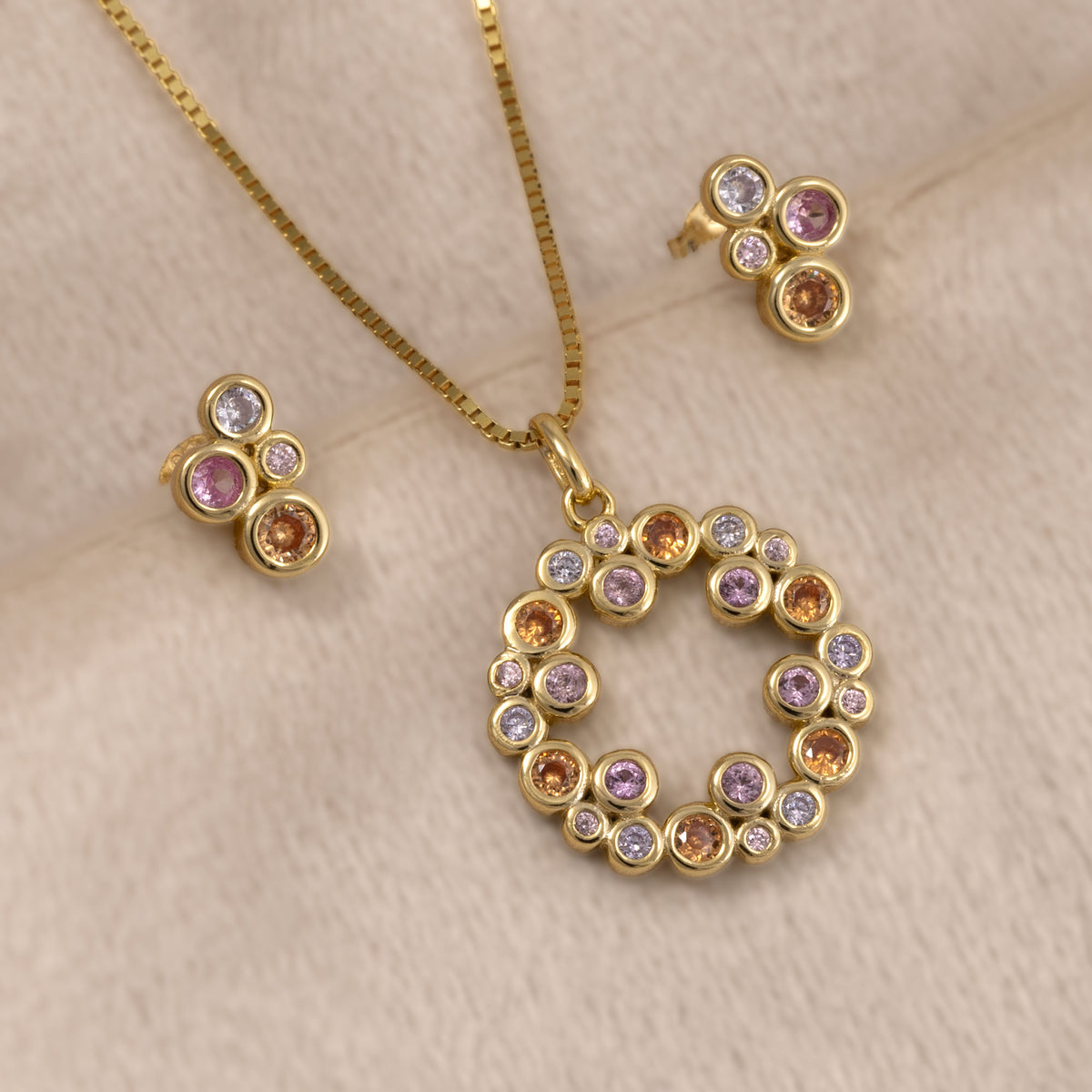 Pair of gold vermeil stud earrings and circular pendant featuring colourful pink, purple, and orange bubble-shaped gemstones in a sculptural cluster design by Azendi. Placed on a cream velvet cloth.