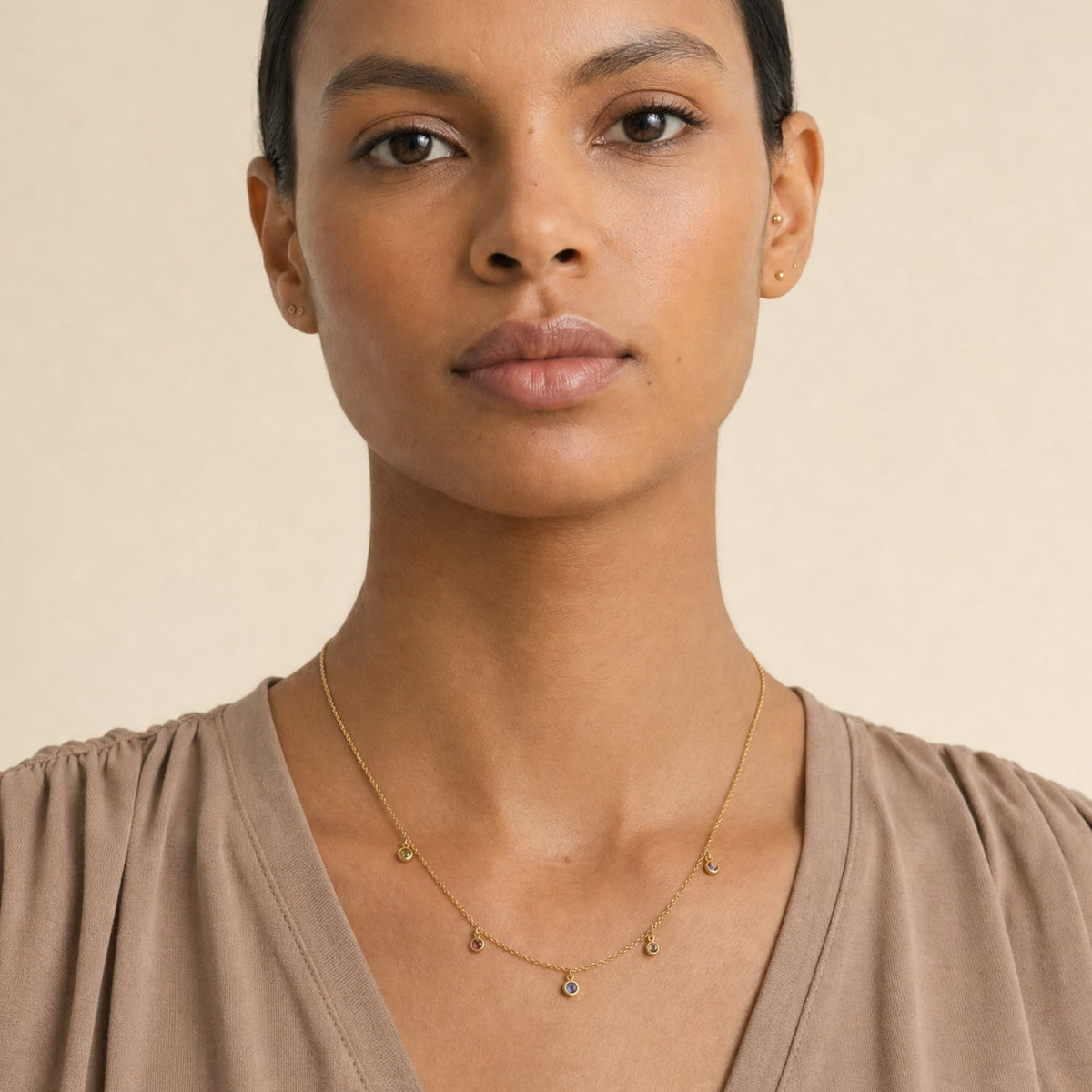 A close-up of a model wearing a delicate gold vermeil chain necklace with small dangling charms, paired with a taupe-brown top against a warm beige background.