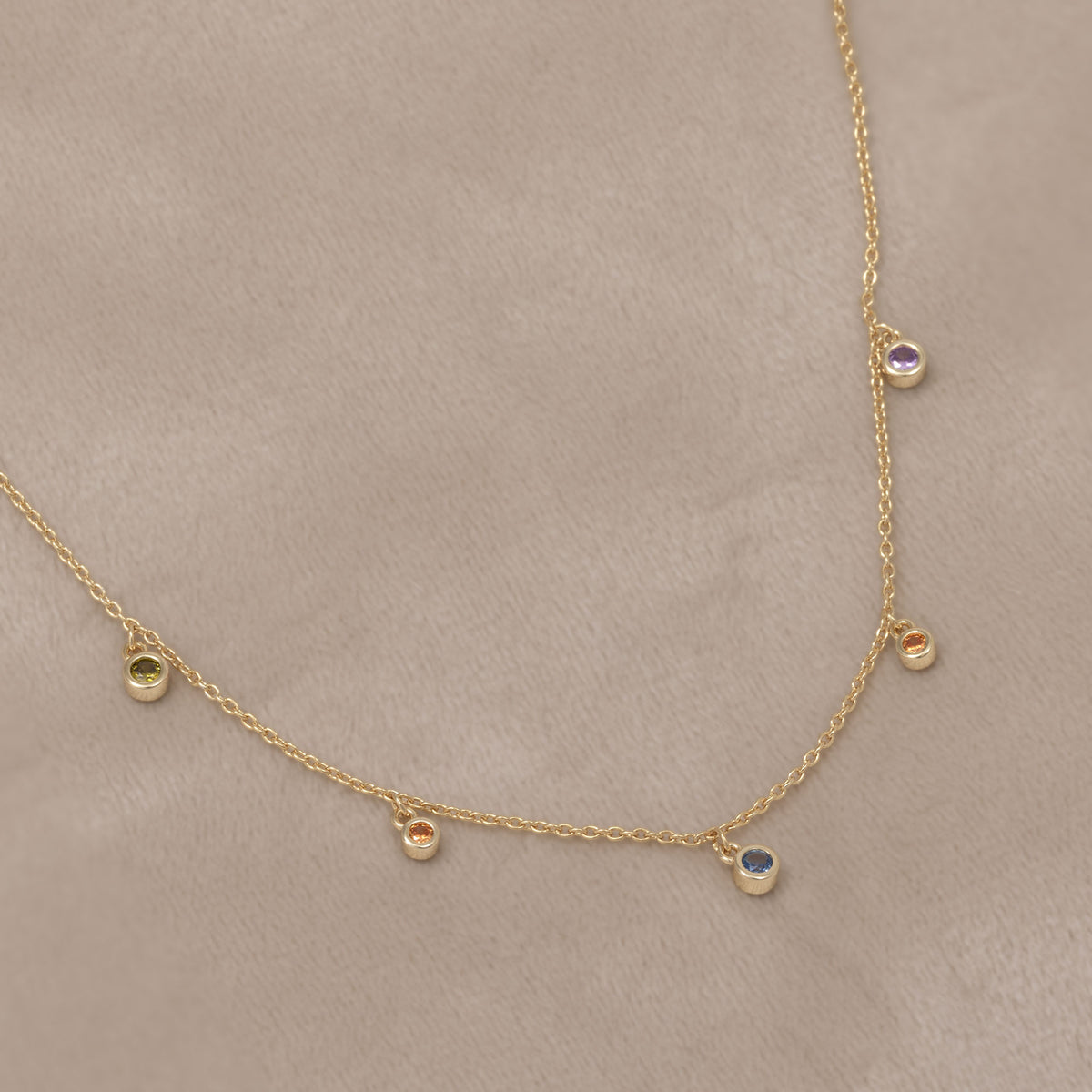 A gold vermeil delicate chain necklace featuring clusters of colourful purple and green bubble-shaped gemstones. Placed on a cream velvet cloth.
