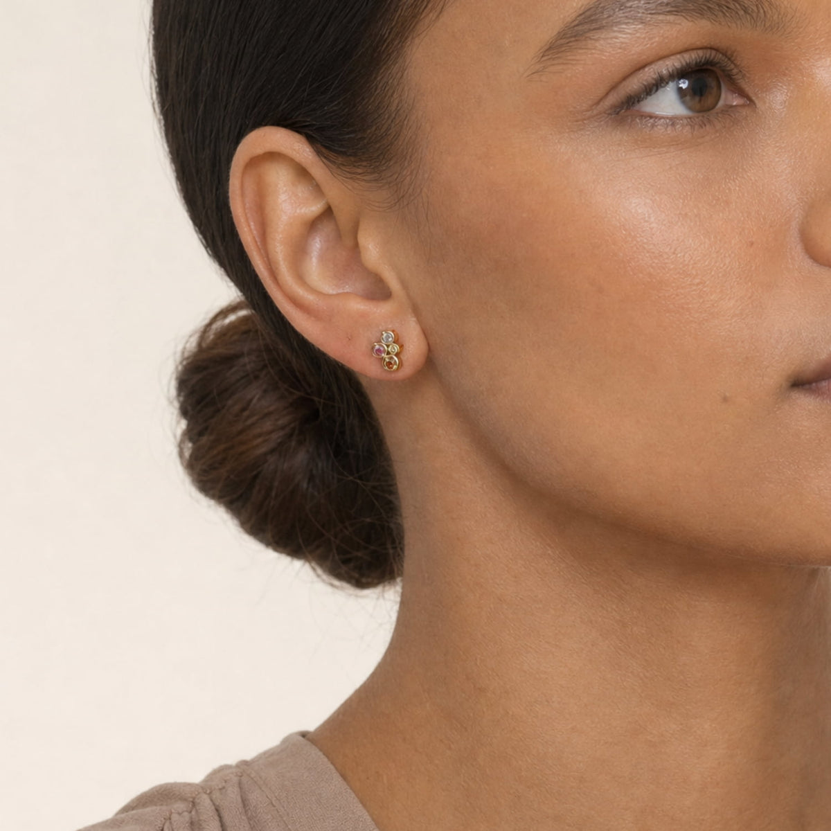 A close-up of a woman's ear wearing a small gold vermeil stud earring with cubic zirconia stones, set against a plain light beige background.