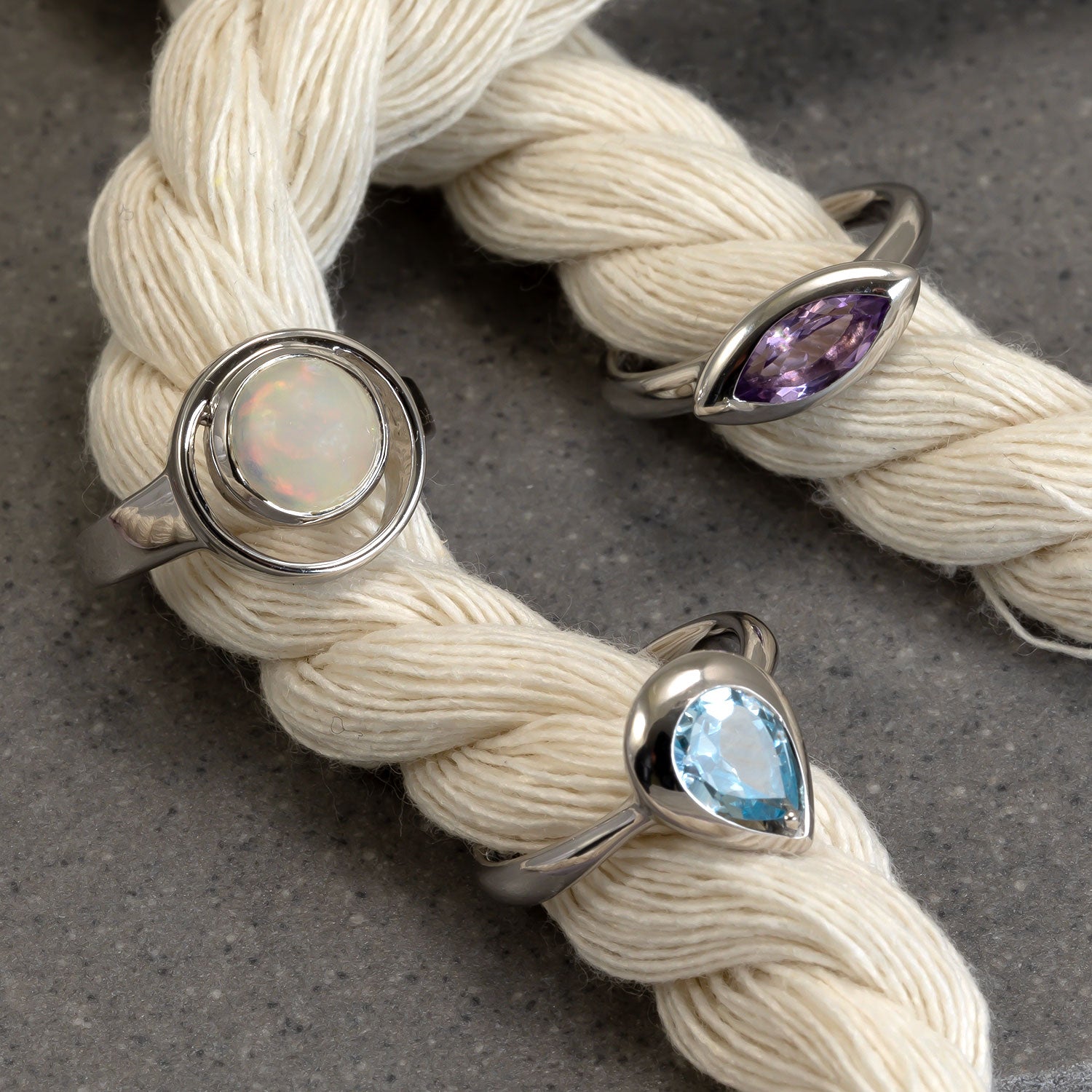 Three sterling silver rings featuring distinct gemstones: a round opal set within a circular silver frame, a marquise-cut amethyst in a sleek bezel, and a teardrop-shaped sky blue topaz with a polished silver band, displayed on intertwined natural cream yarn against a textured grey background.