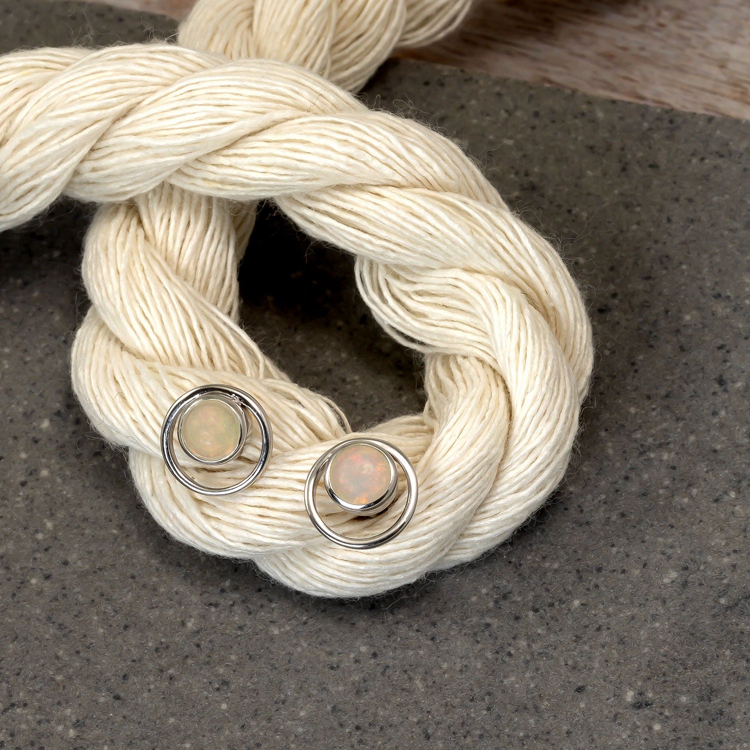 Pair of sterling silver stud earrings featuring circular opal stones set within fine silver hoops, presented on a textured neutral textile backdrop.