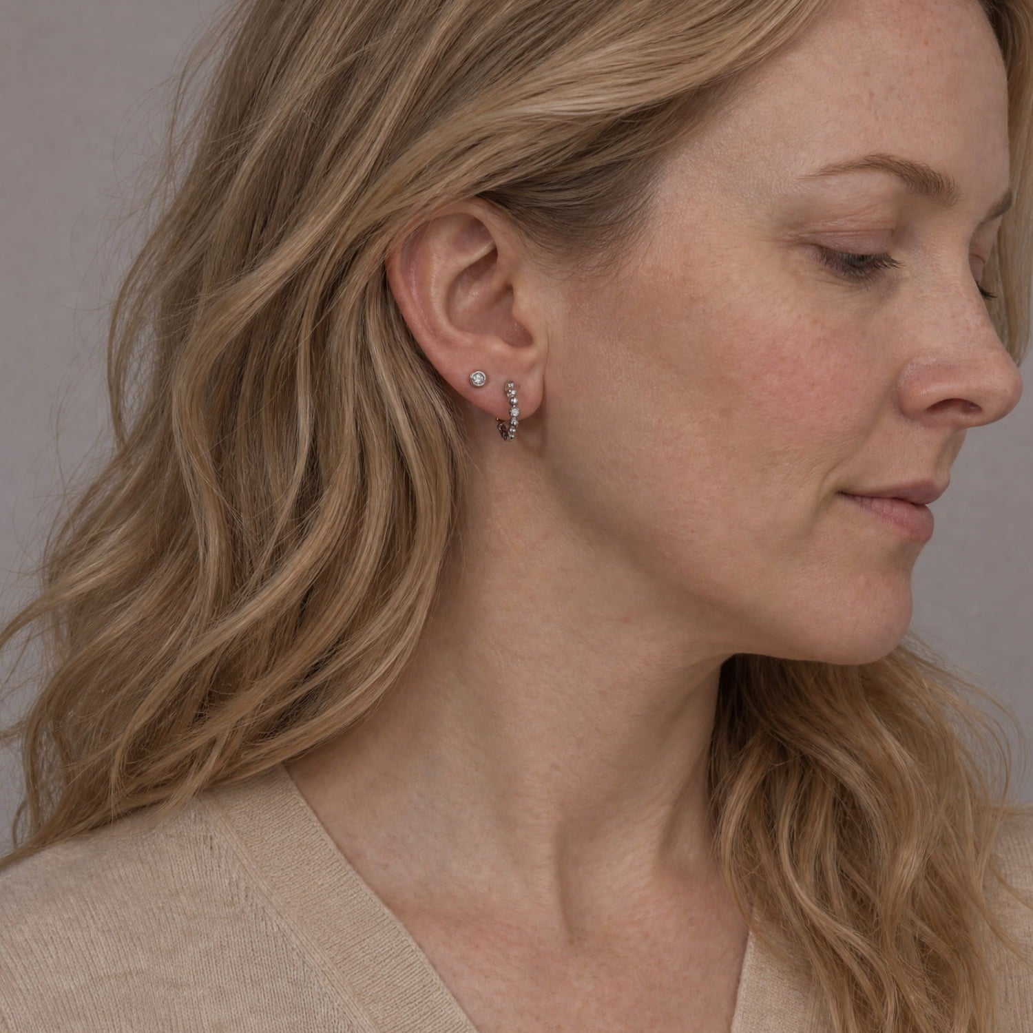 A close-up side profile of a woman with blonde hair wearing a small silver stud earring with a beaded hoop.