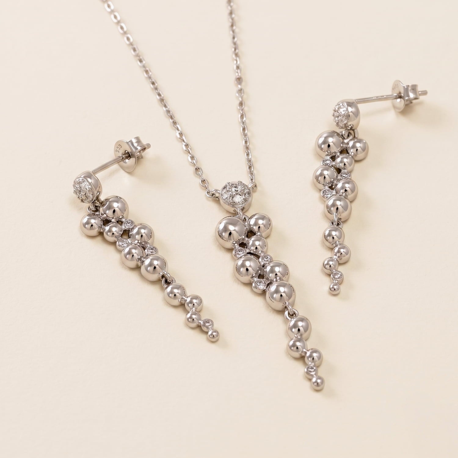 Silver jewellery set featuring a necklace and matching drop earrings with clustered spherical beads on a delicate chain, displayed on a warm cream background.