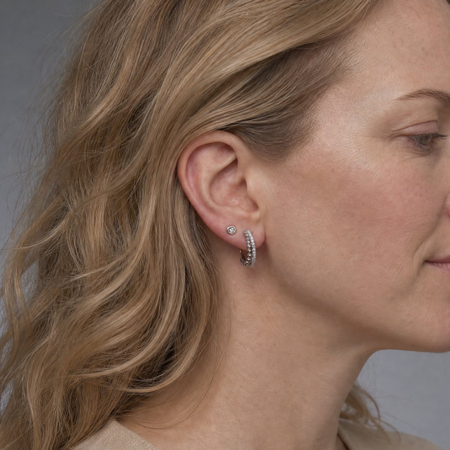 A close-up side profile of a woman with blonde hair wearing a silver hoop earring with clear stones and a matching stud earring, against a soft gray background.
