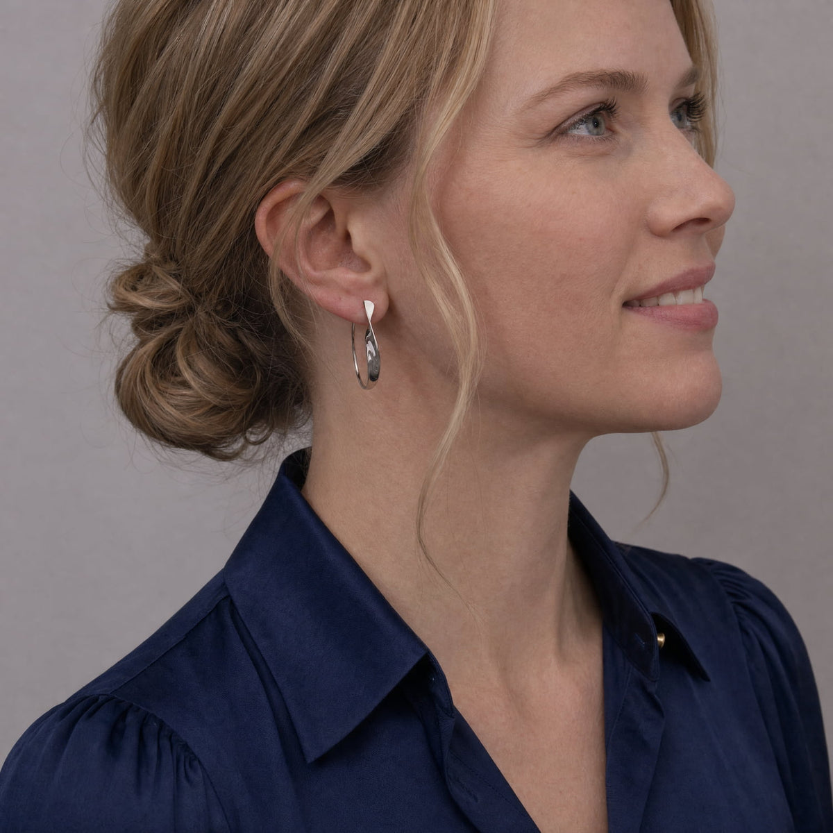 A close-up side profile of a woman with blonde hair in a bun, wearing a silver twisting hoop earring and a navy blue collared blouse against a plain grey background.