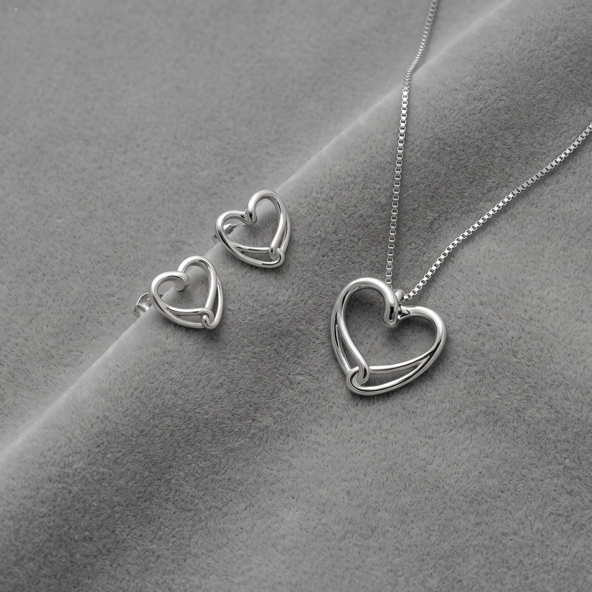 Silver heart-shaped pendant necklace and matching stud earrings on a textured gray fabric background.