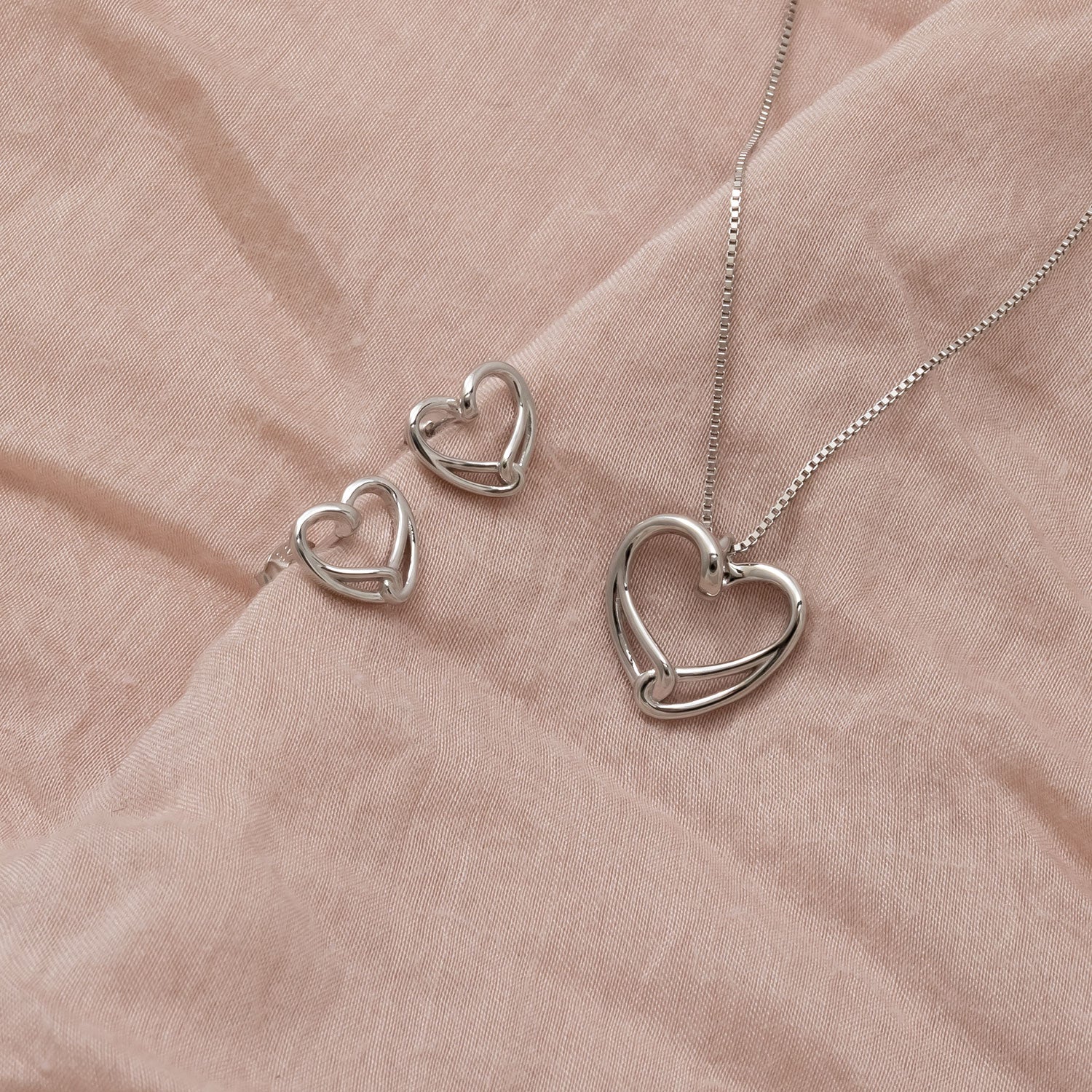 Pair of sterling silver stud earrings featuring an intertwined heart design displayed on a soft neutral fabric alongside a matching pendant necklace.