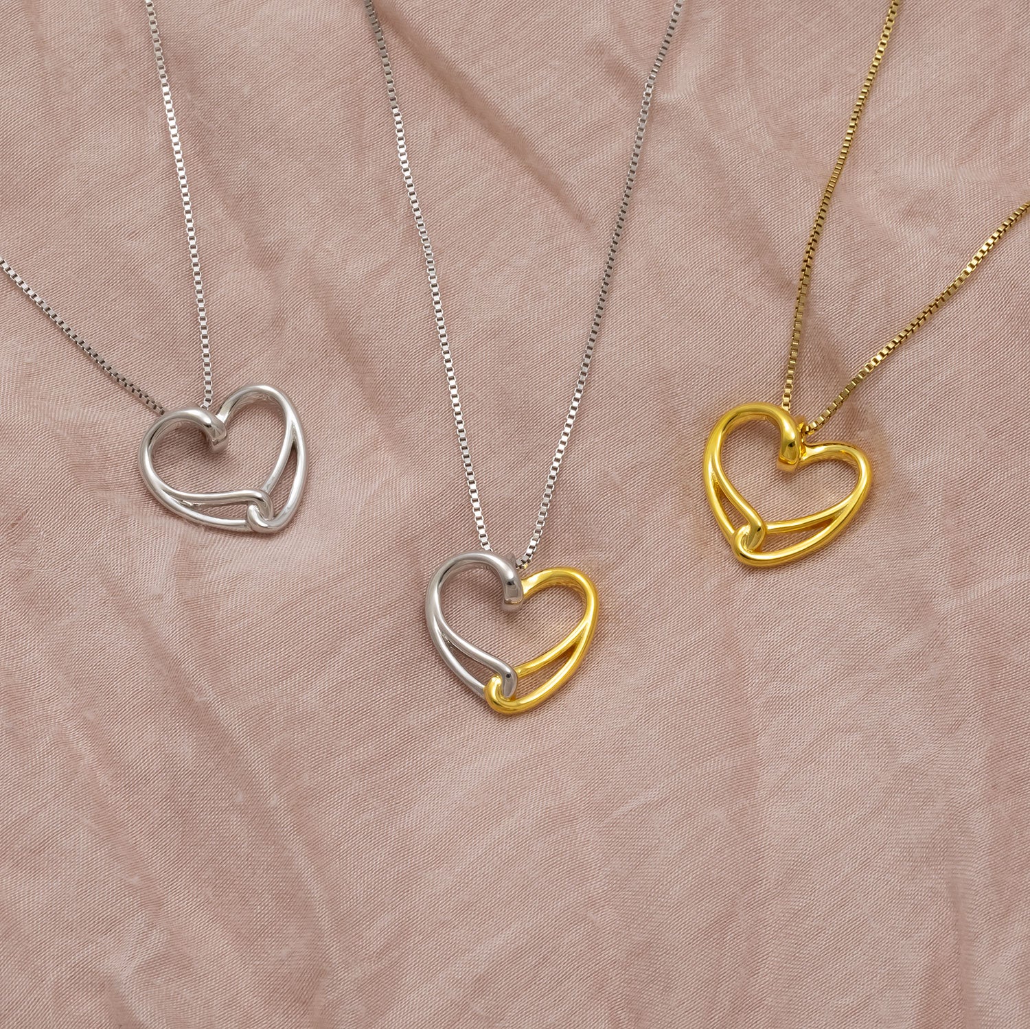 Three sculptural heart pendants on fine chains, featuring sterling silver and gold vermeil intertwined designs, displayed on soft neutral fabric background.